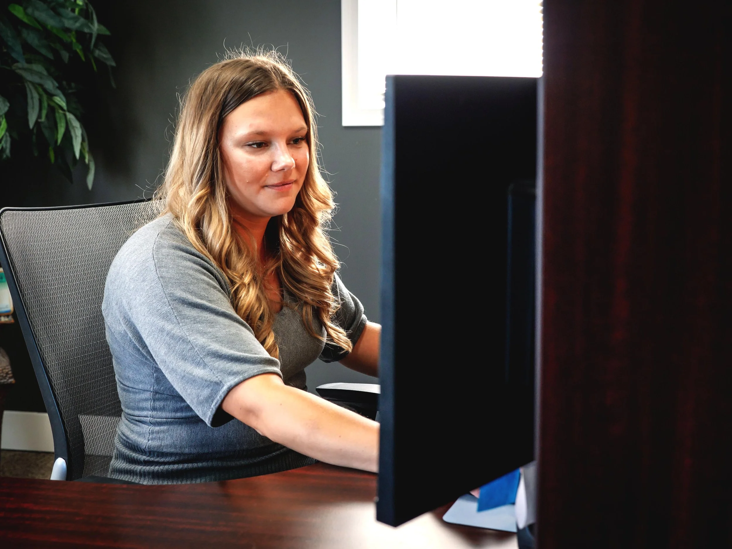 Woman with long wavy hair working at a desk in front of a computer monitor in an office.