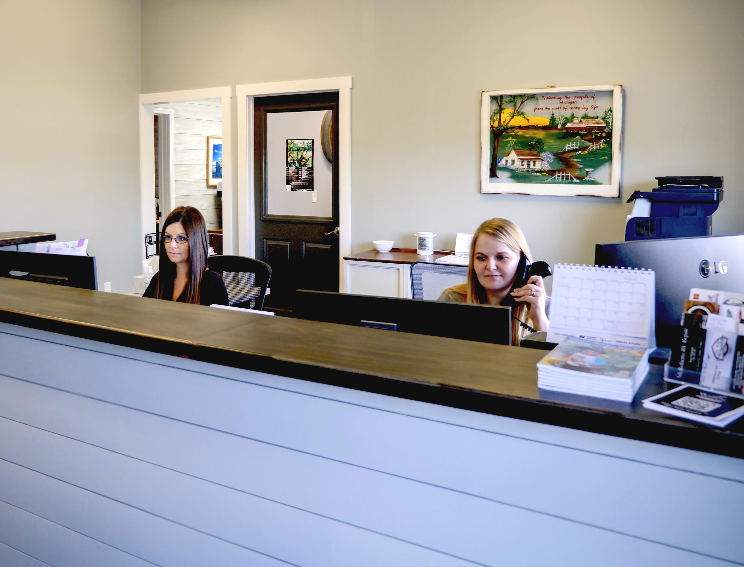 Two women working at a reception desk in an office, one talking on the phone and the other looking at a computer.