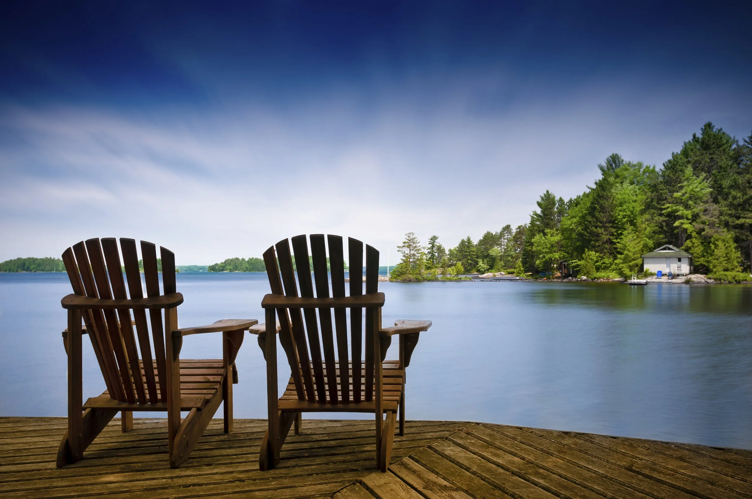 Two wooden Adirondack chairs on a dock overlooking a calm lake with lush green trees on the shoreline and a small house in the distance under a partly cloudy sky.