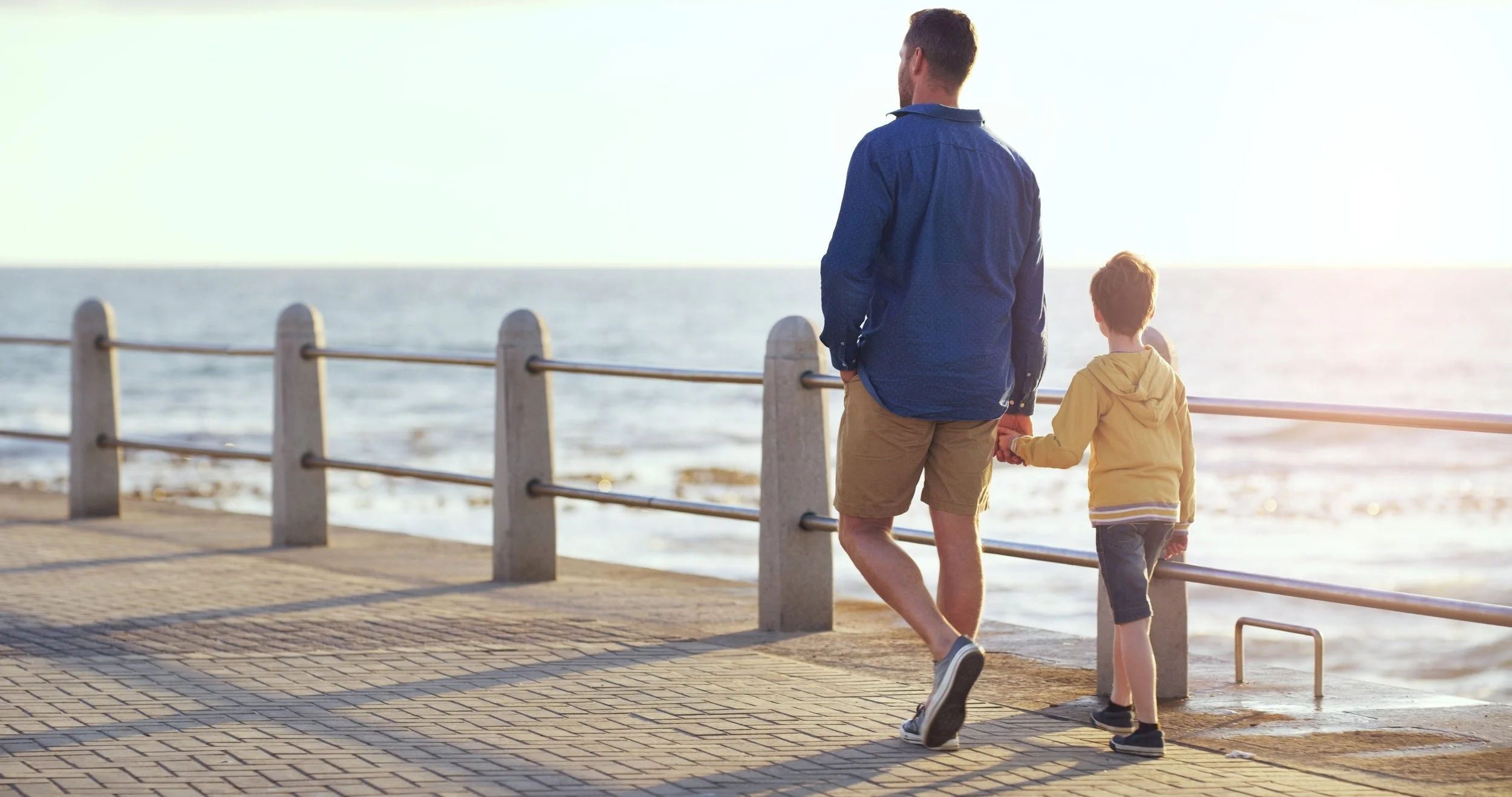 A man and a boy holding hands walking along a seaside promenade at sunset, with the ocean and sky in the background.