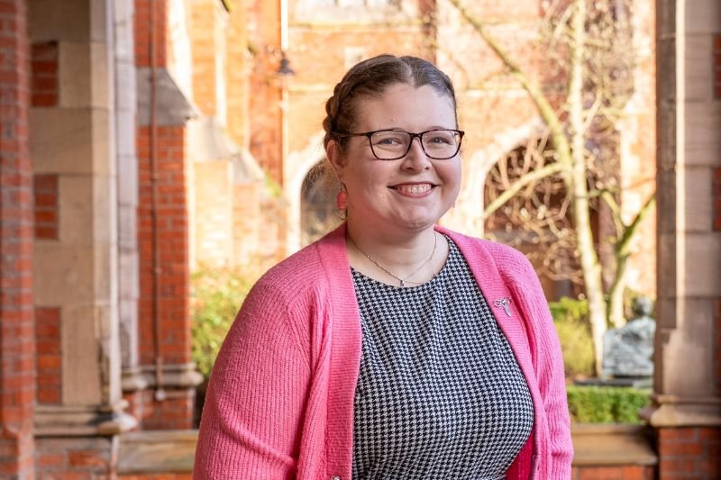 A smiling woman with glasses, wearing a pink cardigan over a patterned top, standing outdoors with brick buildings and trees in the background.
