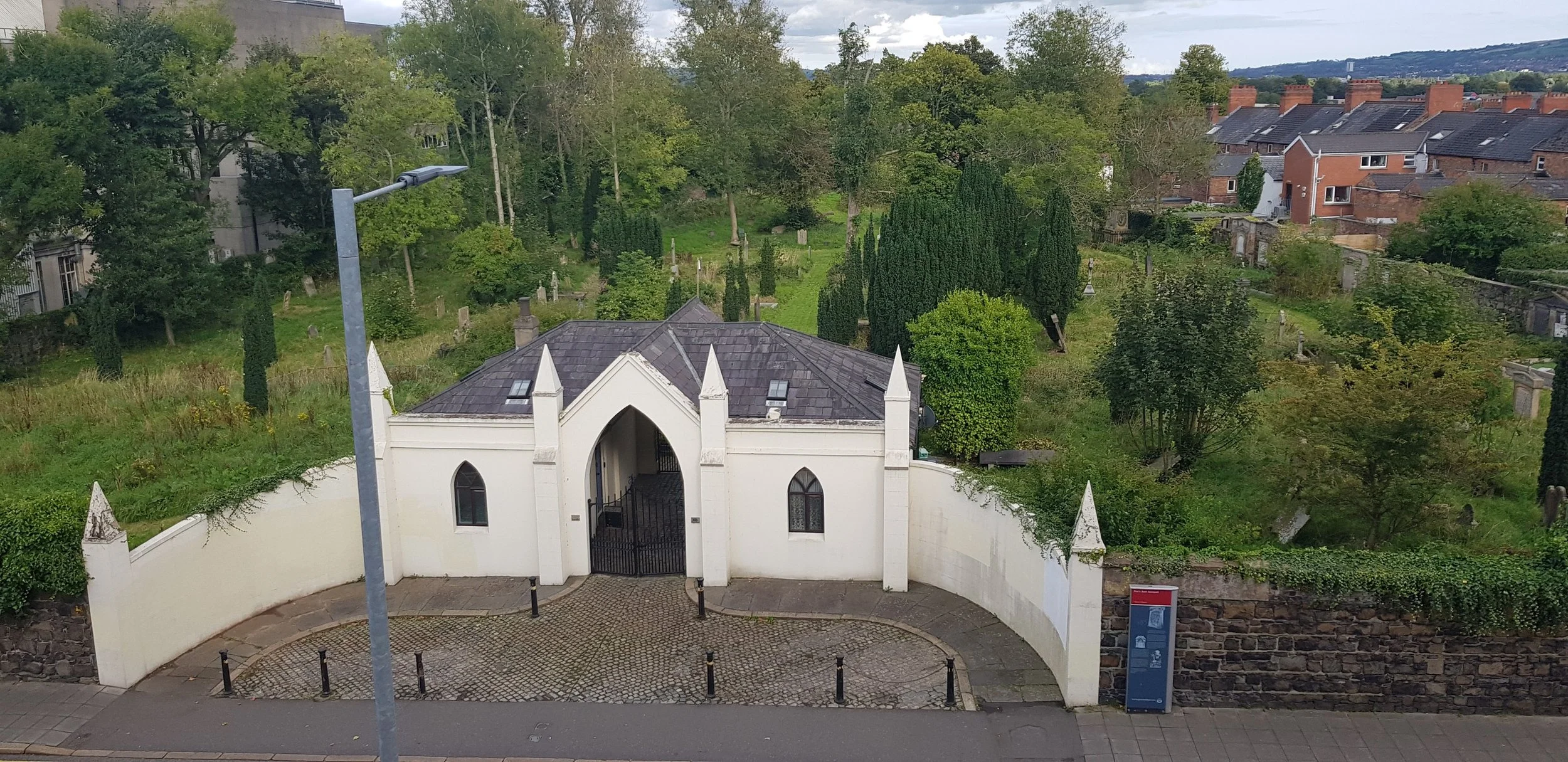 A white Gothic-style building with pointed arches and a gate, surrounded by a curved white wall, located on a street with a lamppost and an information board. Behind it, there is a green area with trees and a background of houses and hills.