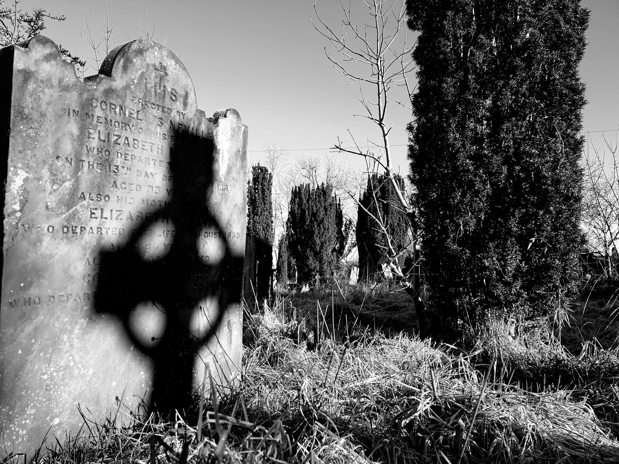 A black and white photograph of the shadow of a celtic cross projected onto a very old gravestone.