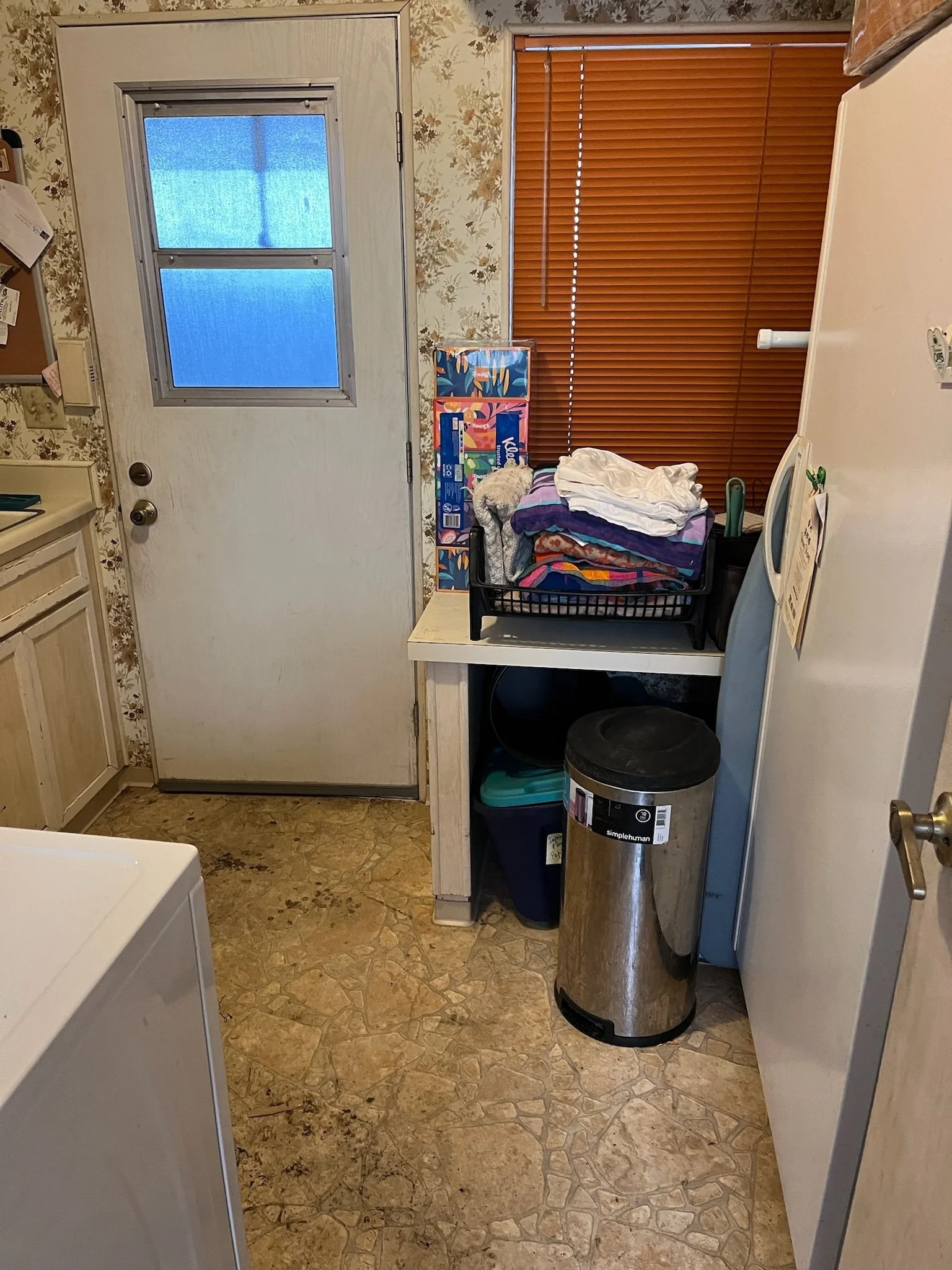 A laundry room with a door, window blinds, and a trash can. There is a basket with folded clothes and other laundry items on a small table, and a washing machine is partially visible in the corner.