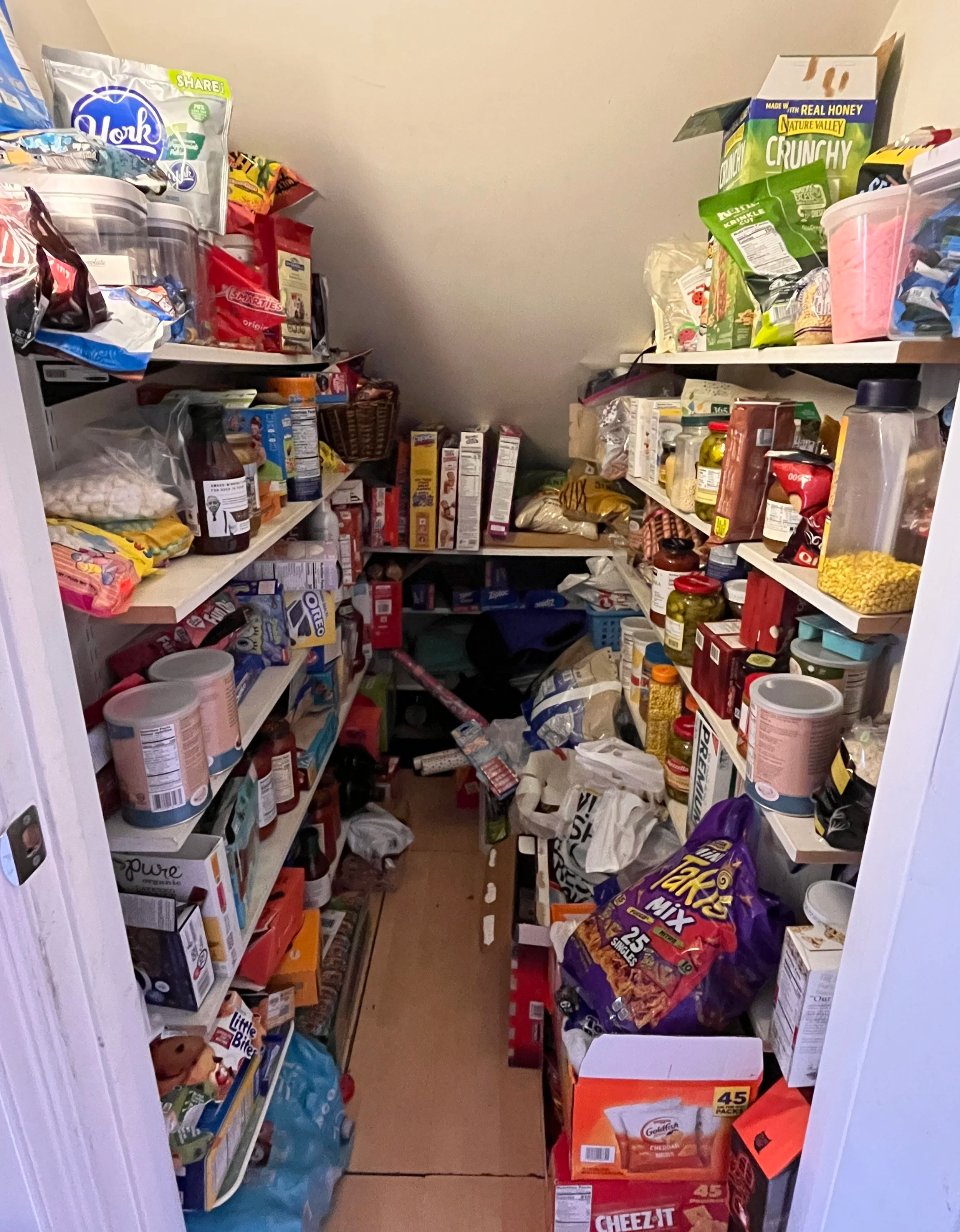 A small pantry filled with shelves of snacks, canned goods, and boxed foods, with some items on the floor.