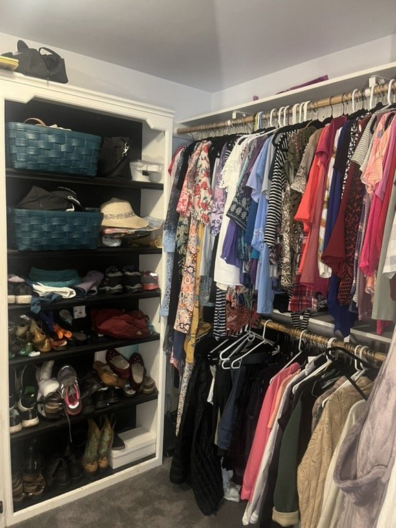 A walk-in closet with shelves on the left filled with shoes, hats, and accessories, and hanging clothes on racks on the right, including various shirts, dresses, and other clothing items.