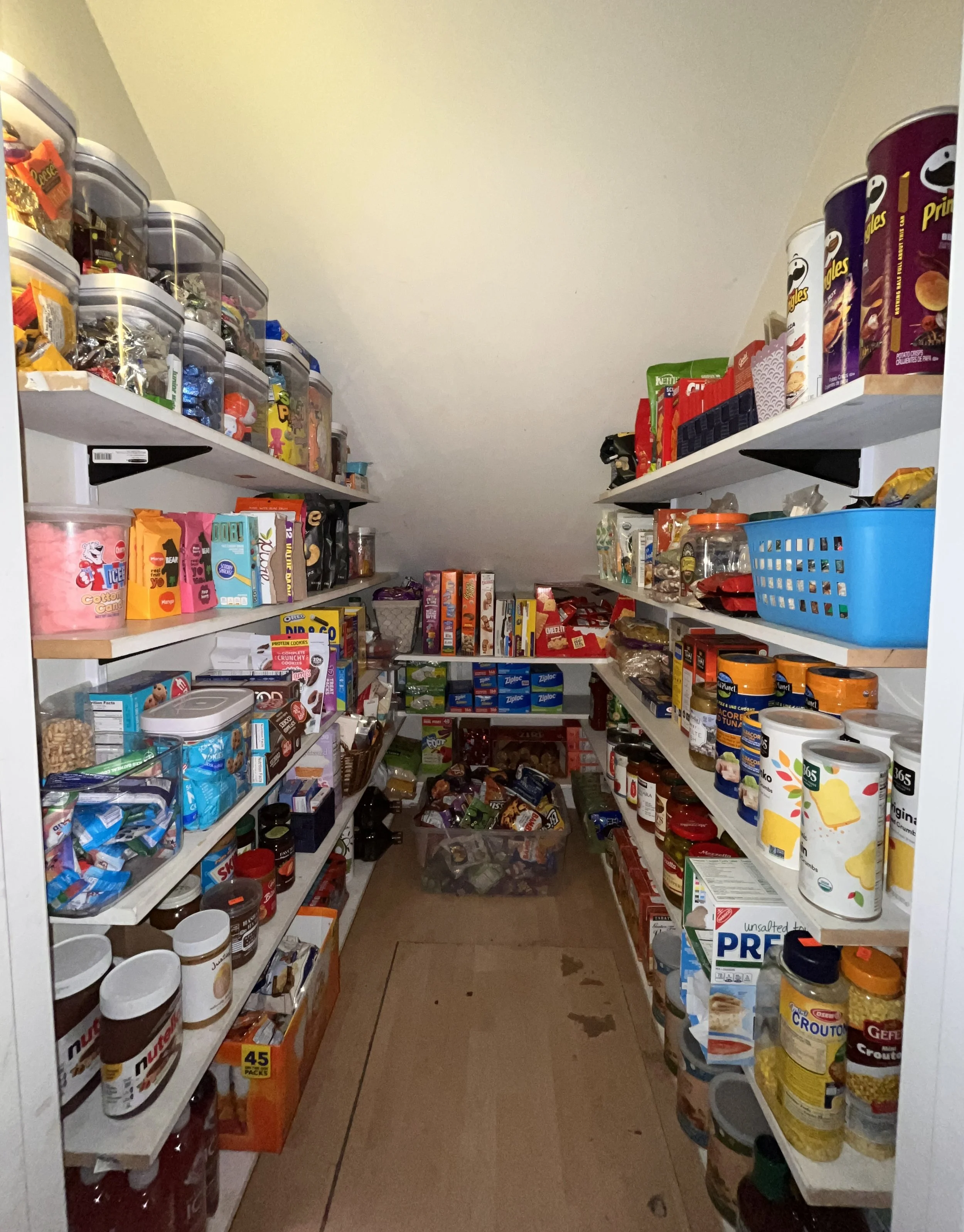A small grocery or pantry closet with white shelves filled with food products, including cans, boxes, and jars, organized on both sides and on the floor.