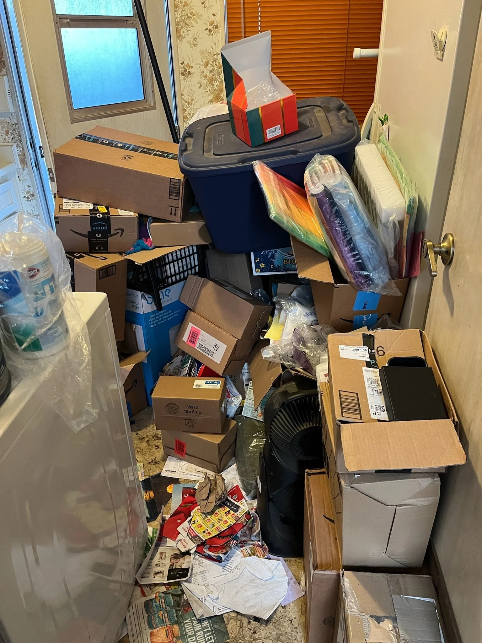 A small cluttered space filled with various boxes, wrapping paper, and miscellaneous household items. There is a window with blinds, and the floor is covered with paper and packaging materials.