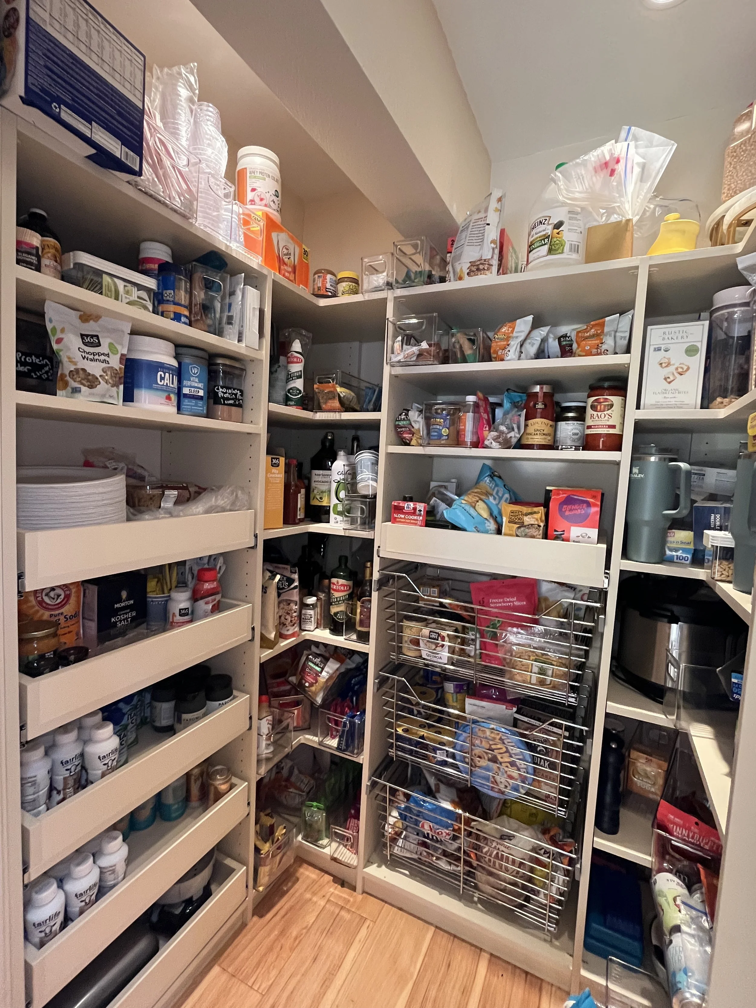 A pantry with white shelves filled with various food and household items, including canned goods, jars, snack packets, and bottles, with a wooden floor.