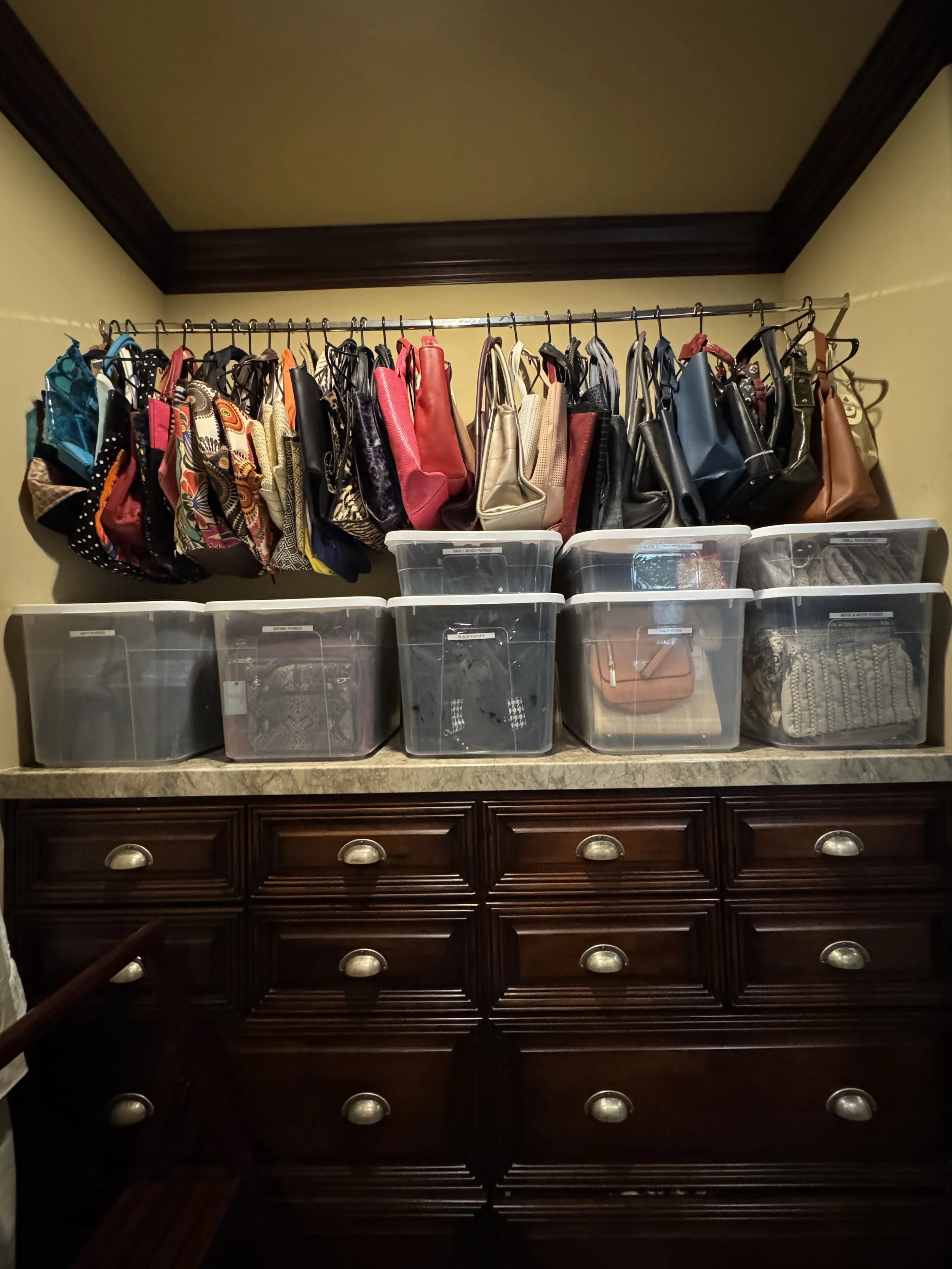 A wooden dresser with three drawers and metal handles, against a beige wall with crown molding. On top of the dresser are transparent plastic storage bins filled with various items, and above them, a hanging rod with numerous handbags and purses of d