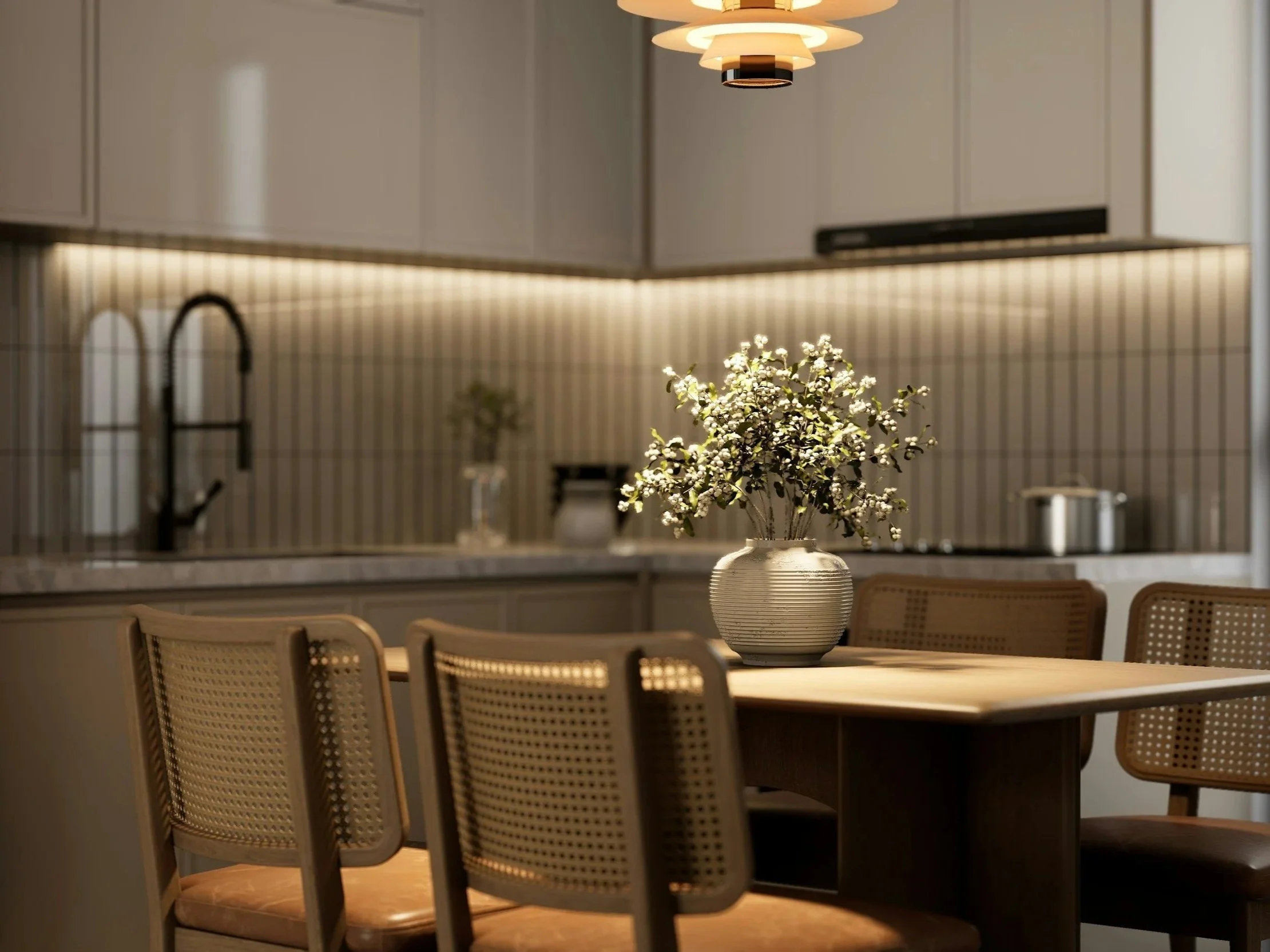 A modern kitchen with beige cabinets, a black kitchen faucet, a beige tiled backsplash with thin vertical lines, and a dining table with woven back chairs. There is a white ceramic vase with white flowers on the table.