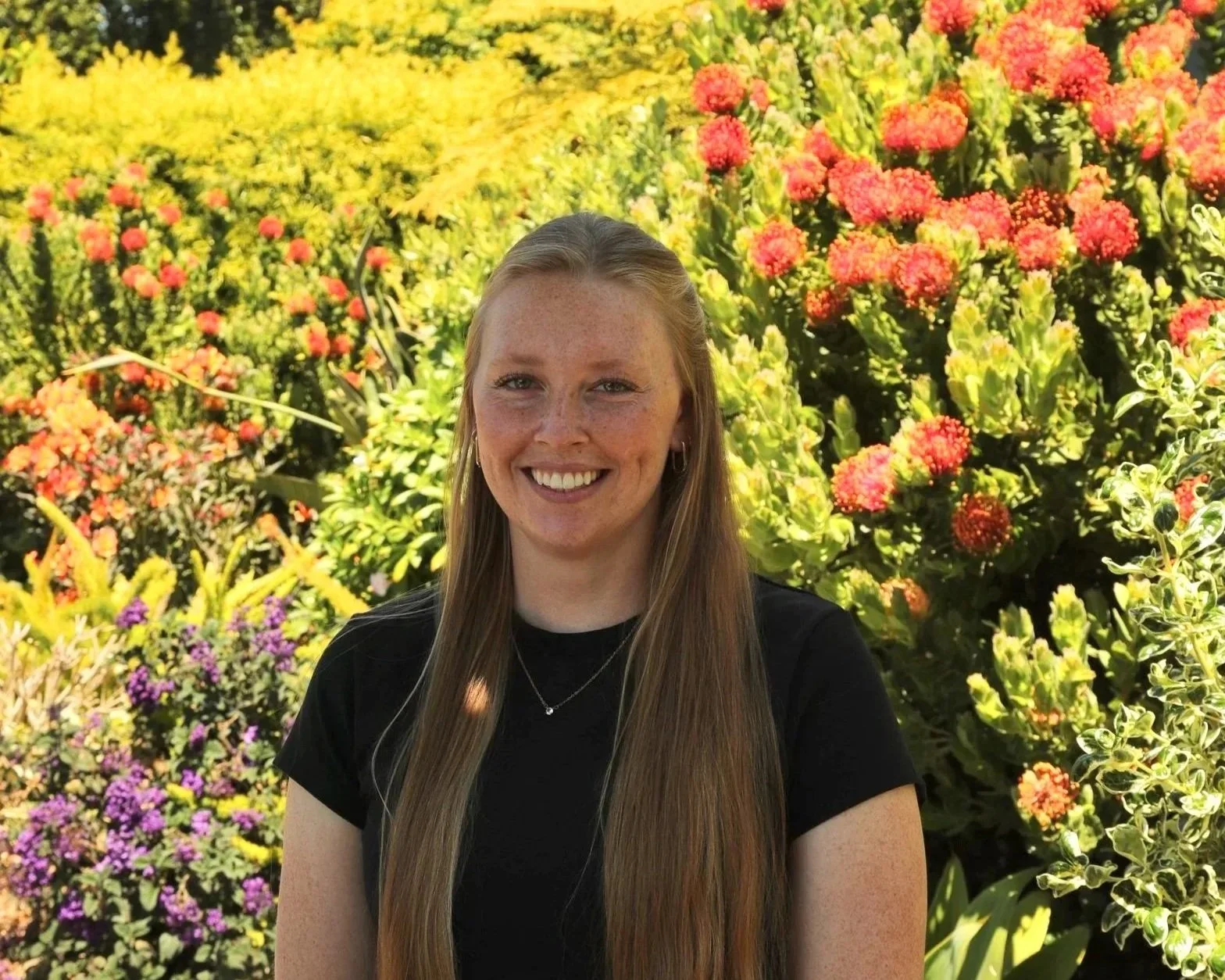 A woman with long blonde hair, wearing a black shirt and a small necklace, smiling in front of colorful flowers, including pink, yellow, and purple blooms.