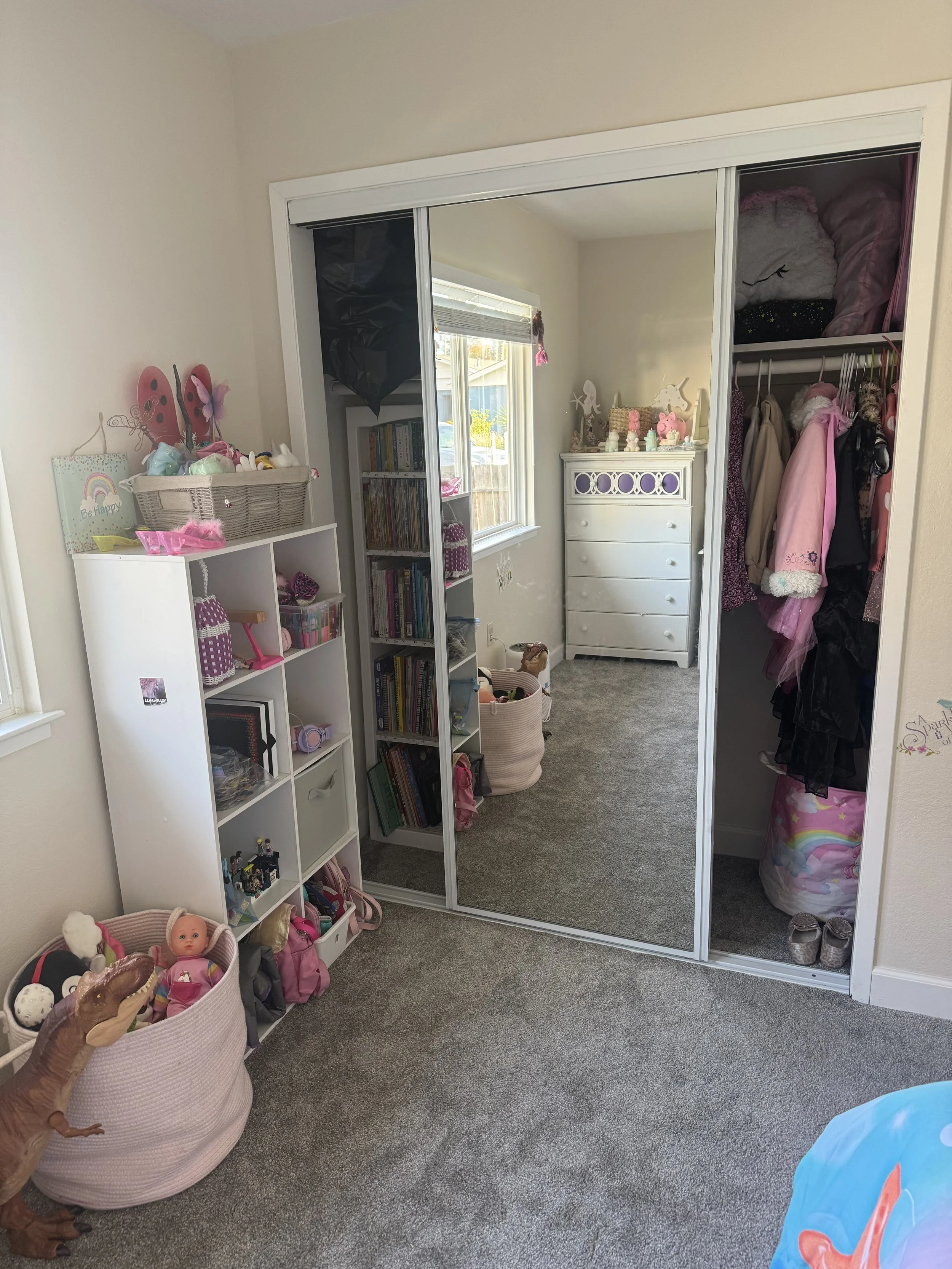 A children's bedroom with a white shelving unit filled with dolls and toys on the left, a pink toy bin with dolls and plush toys in the foreground, and a mirrored closet door reflecting a window and a white dresser decorated with toys and figurines o