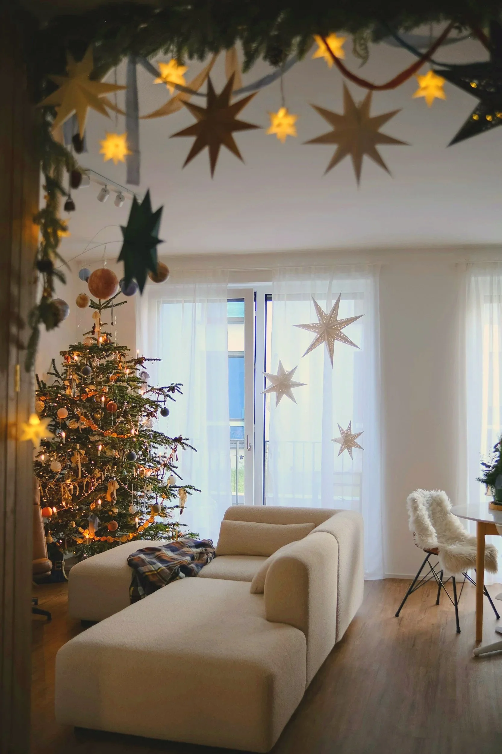 Living room decorated for Christmas with a Christmas tree, star-shaped hanging ornaments, and white curtains on a large window.