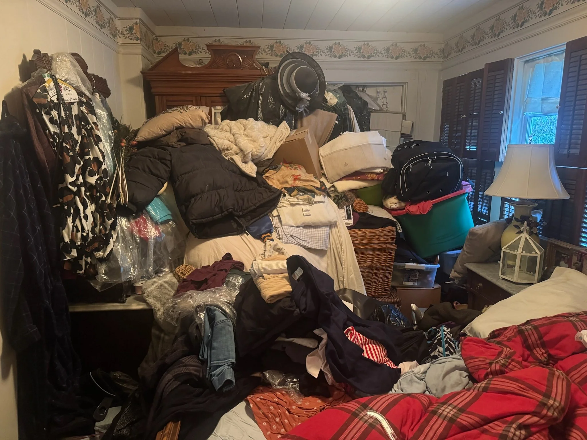 A cluttered bedroom filled with clothes, pillows, and various items piled on furniture and the floor, with a window with partially open wooden shutters on the right and a dresser in the background.
