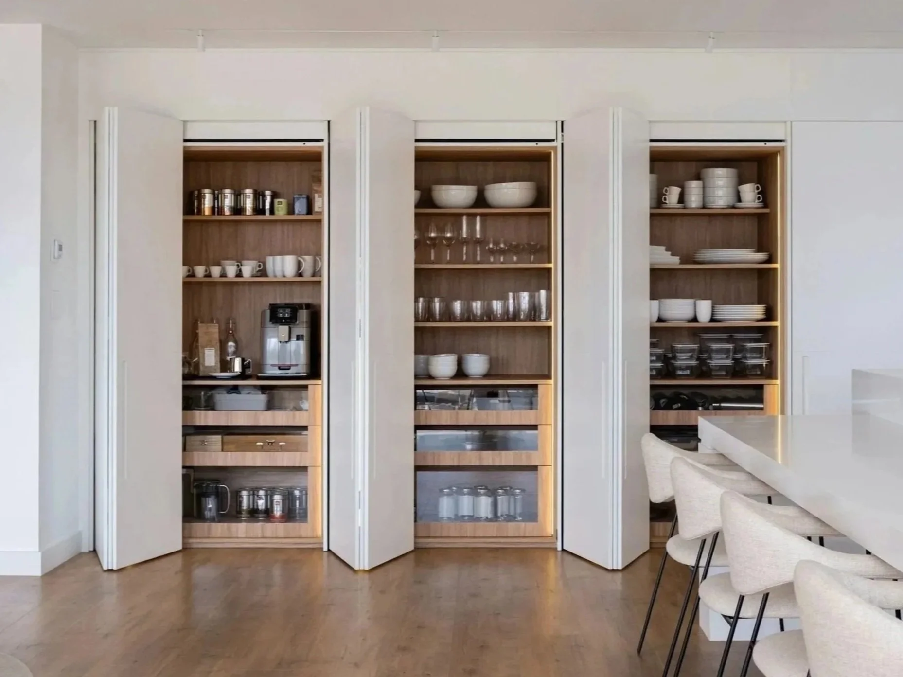 Kitchen cabinetry with open shelves filled with white bowls, cups, glasses, and various kitchen items. Part of a white kitchen island with chairs on the right side.