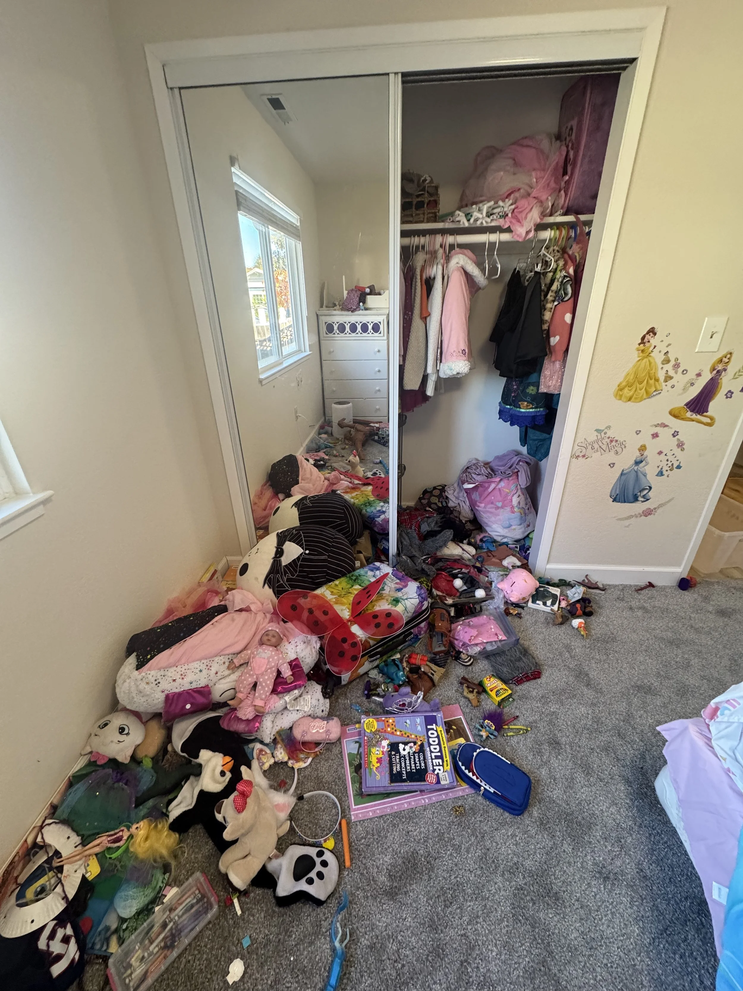 Disorganized bedroom closet with clothes hanging, stuffed animals, toys, and miscellaneous items scattered on the carpet, including a children's toy set and decorations on the wall.
