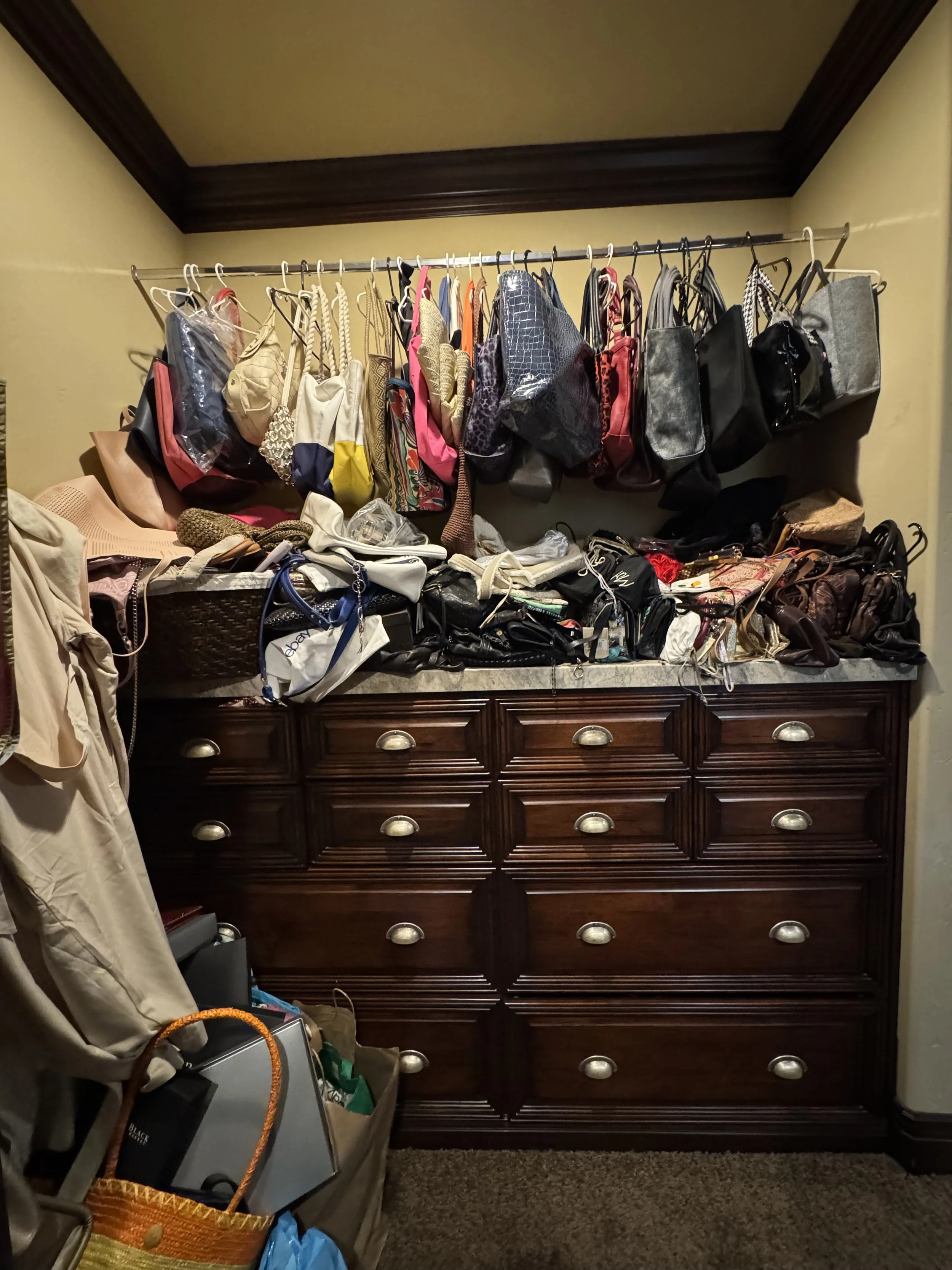 A wooden dresser filled with clutter including bags, purses, and various items. An upper rack contains hanging bags and purses, some in plastic covers. The lower surface and nearby area hold more bags, papers, and miscellaneous objects. The room has 
