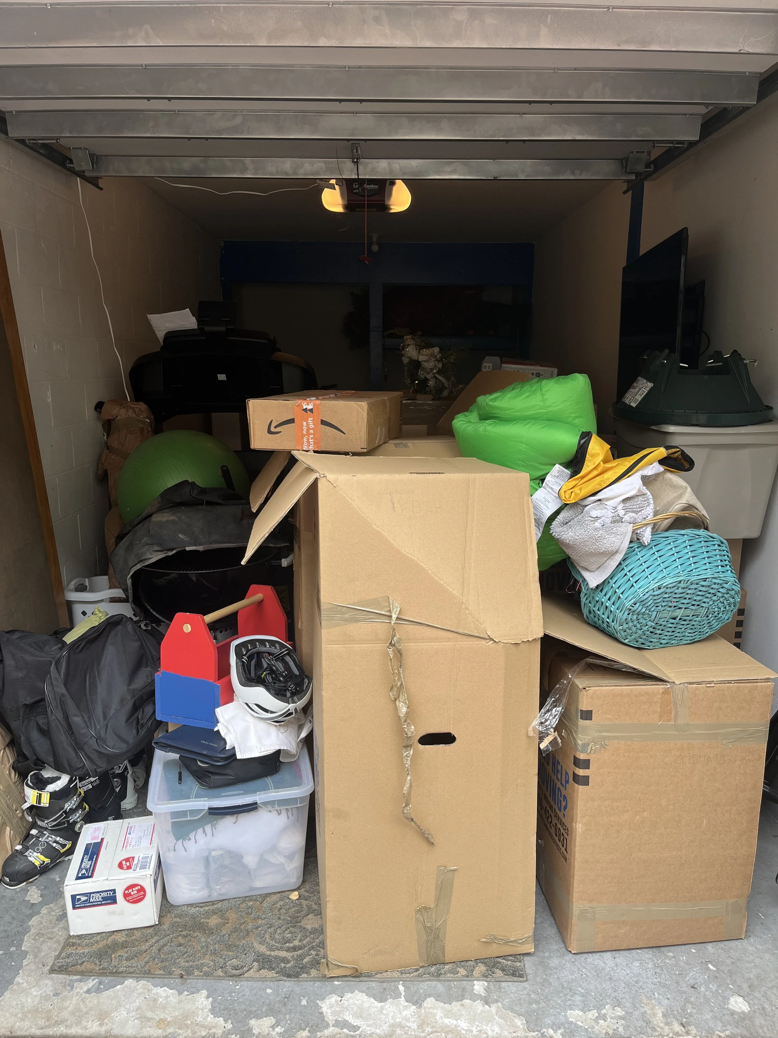 A cluttered garage with cardboard boxes, a green exercise ball, a helmet, plastic storage containers, and various household items.