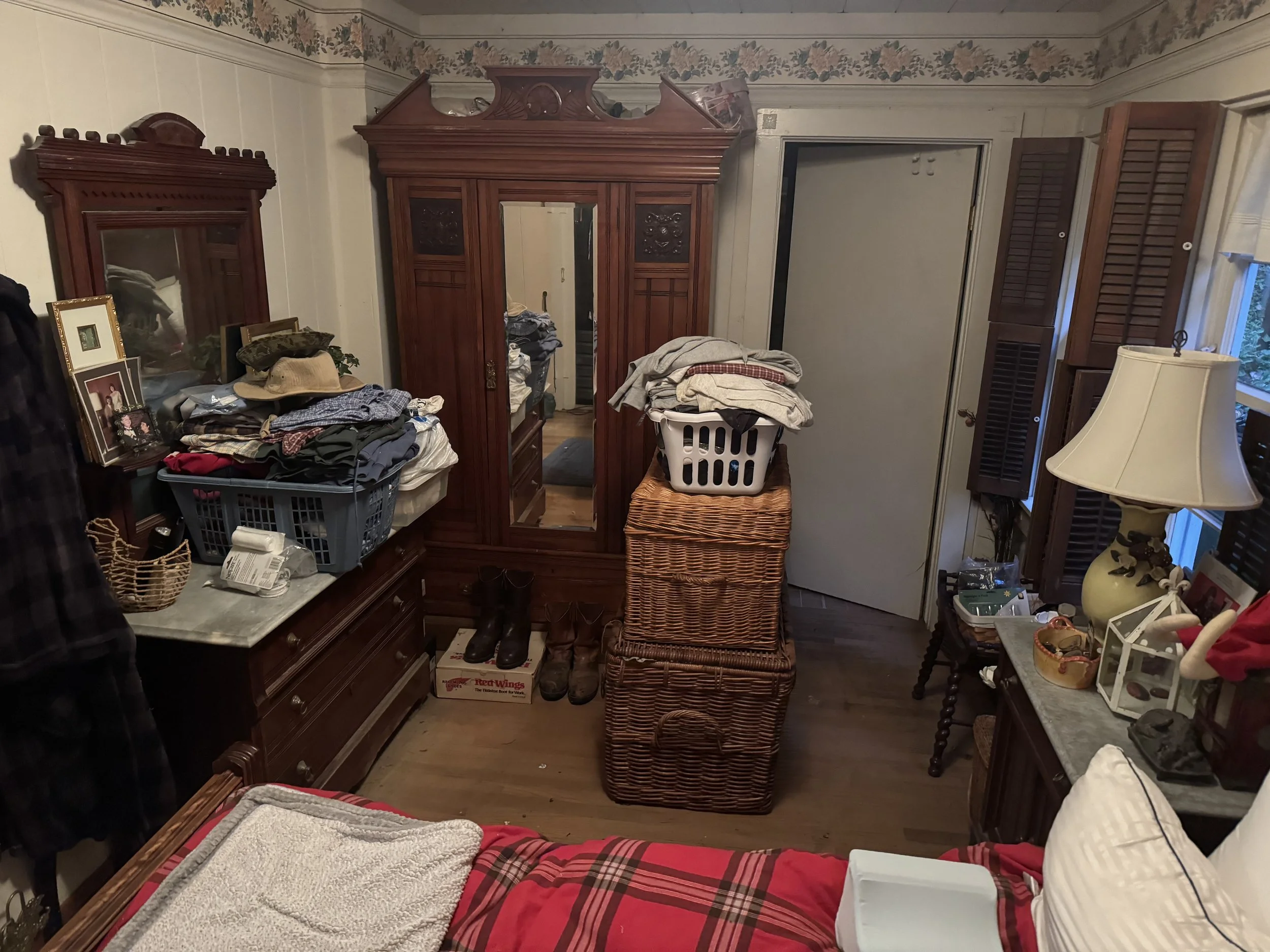 A bedroom with furniture, clothes, and various household items, including a large wooden wardrobe, baskets, a bed with a plaid blanket, and shelves with decorative objects.