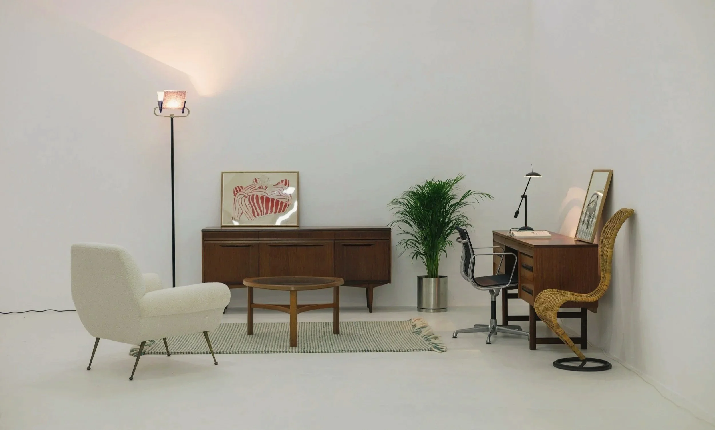 Minimalist living room with white walls, a cream armchair, a wooden coffee table, a decorated sideboard with art, a plant, a mid century modern style desk with framed art, a chair, and a lamp.
