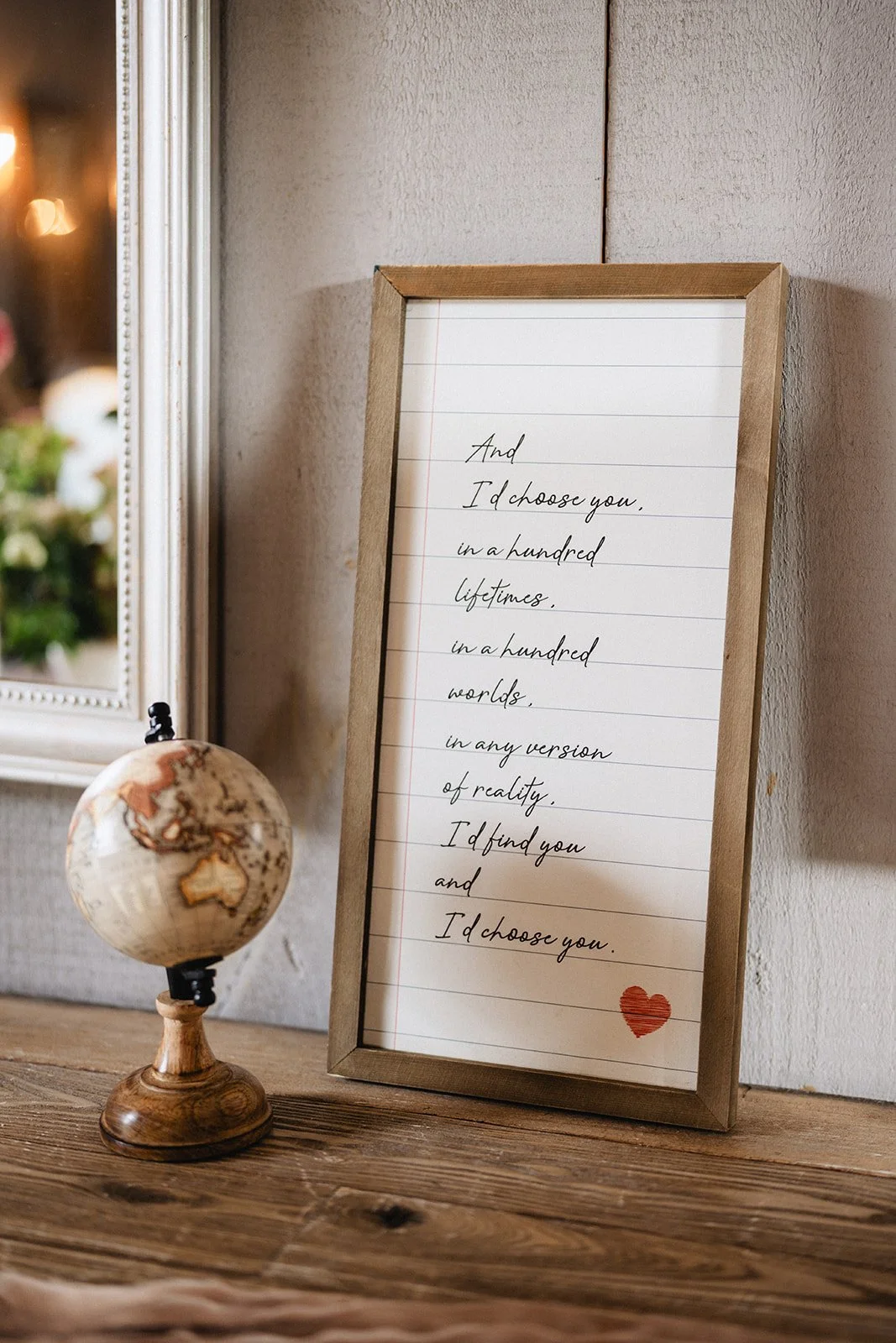 A framed handwritten quote on lined paper with a small red heart at the bottom right, placed on a wooden surface with a small globe beside it, in a cozy indoor setting.