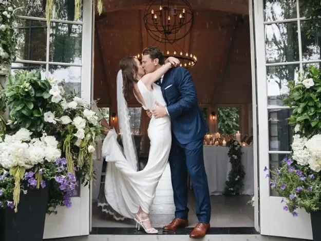 A couple in wedding attire sharing a kiss inside a decorated venue with floral arrangements and large windows.