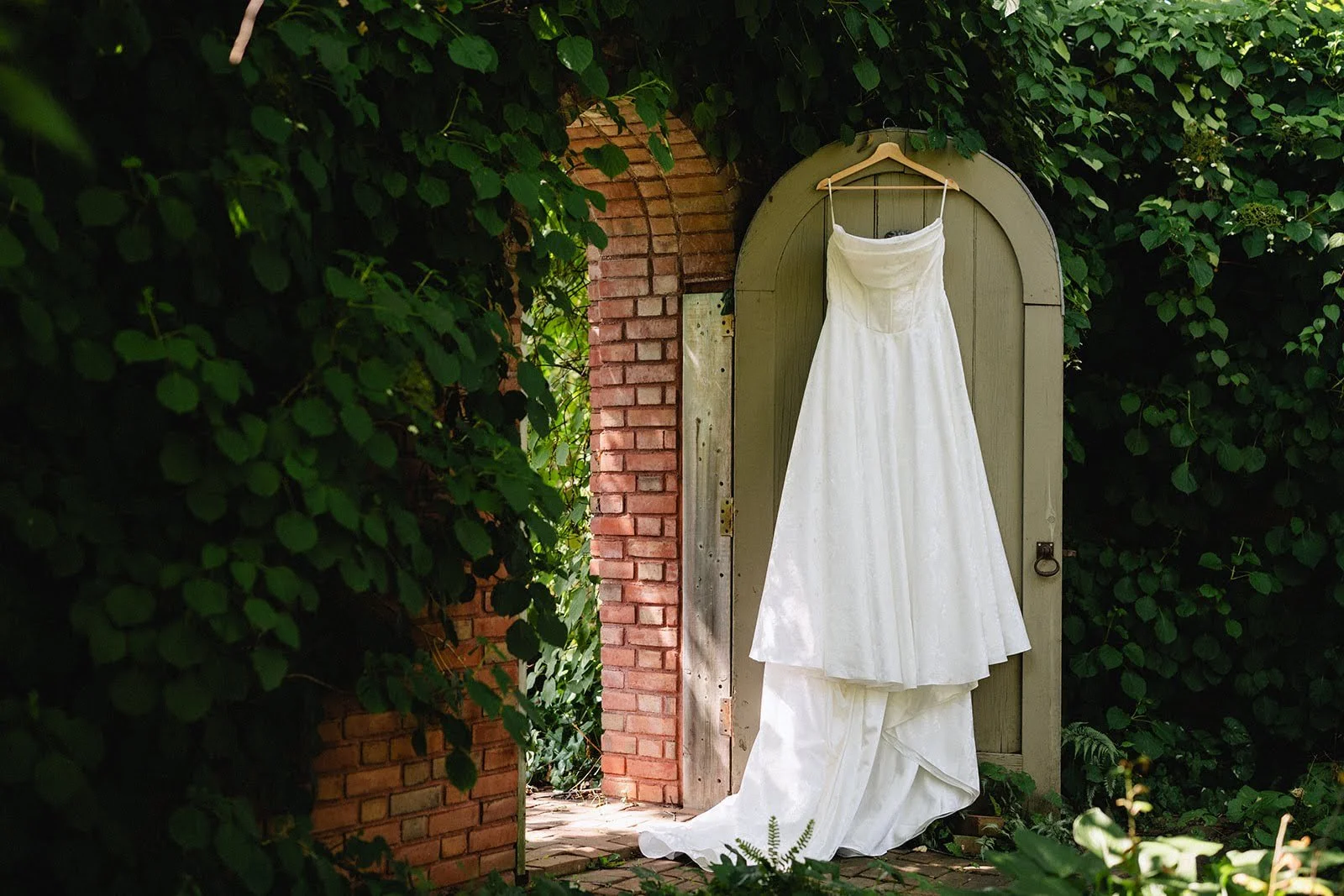 A white wedding dress hangs on a hanger on a wooden door, set against a backdrop of lush greenery and brick archway.