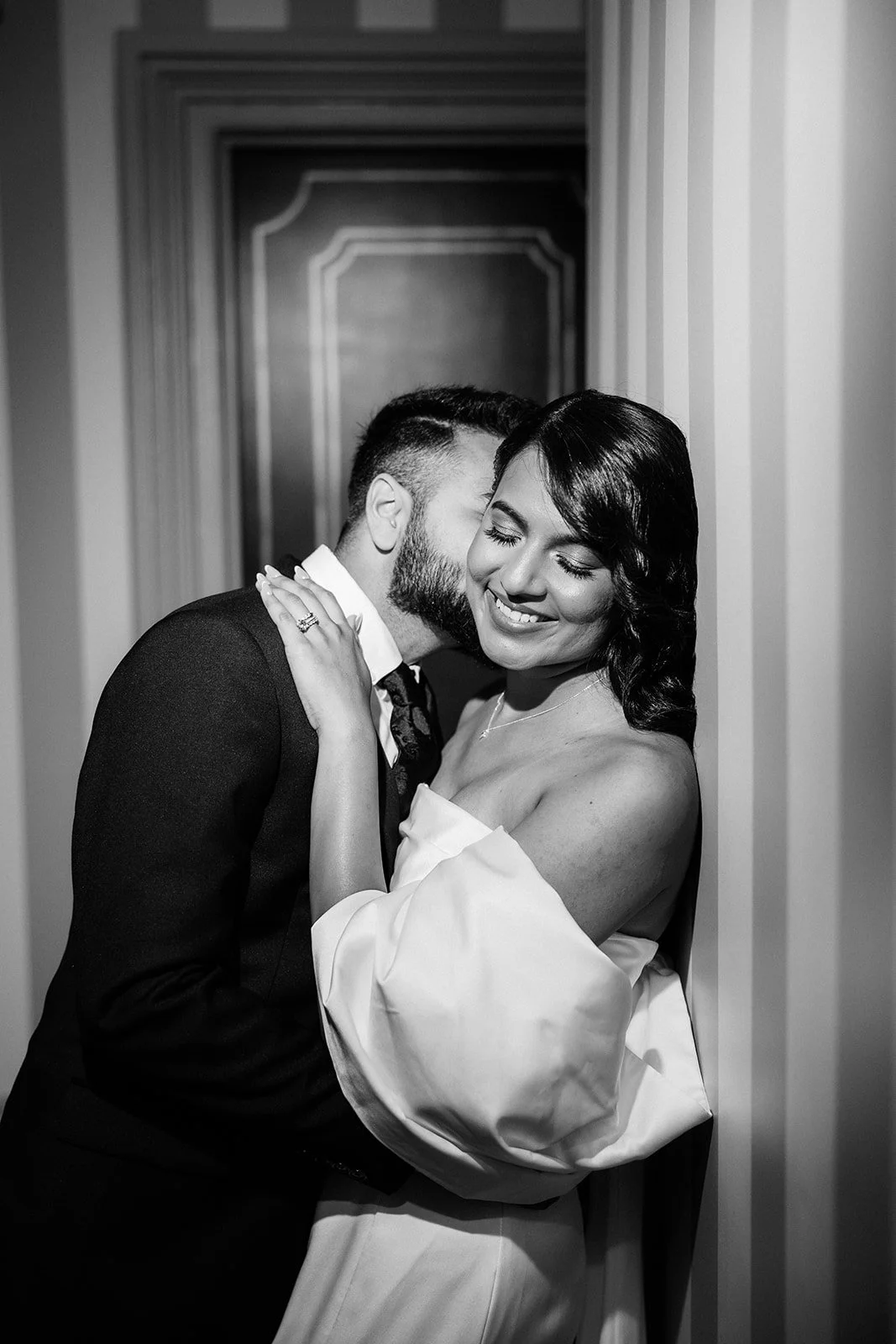 A smiling woman in an off-the-shoulder dress and a man in a suit sharing a tender moment, with the man whispering in the woman's ear in a narrow hallway.