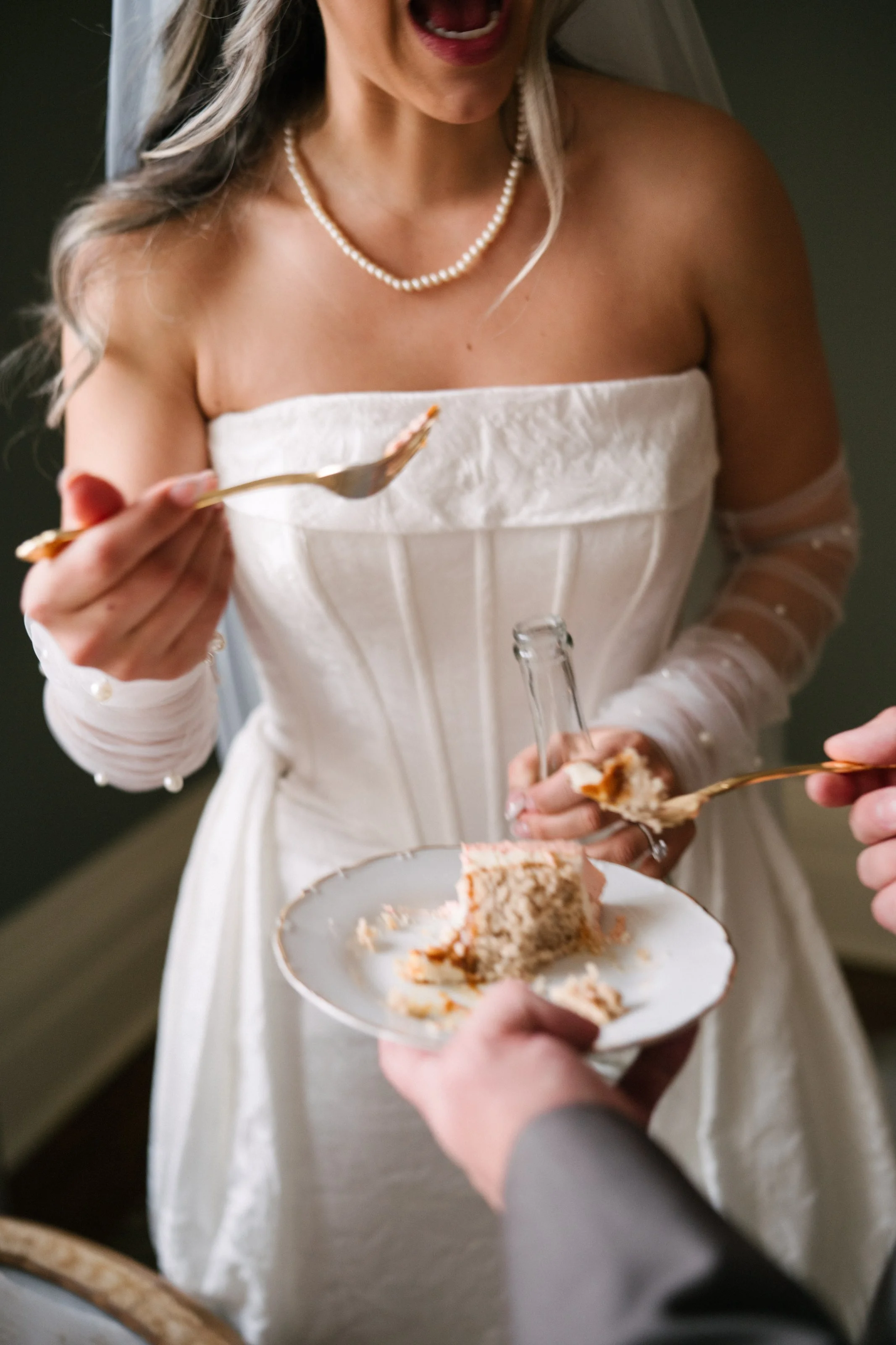 Bride in wedding dress and pearl necklace taking a bite of wedding cake with a fork while someone hands her a piece of cake on a plate.
