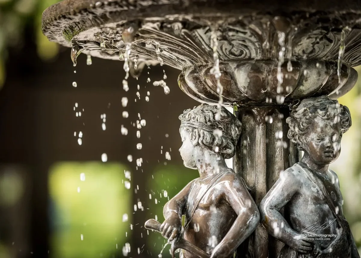 Close-up of a decorative fountain with water flowing from the top, featuring two bronze-style statues of children back-to-back holding objects, with a blurred outdoor background.