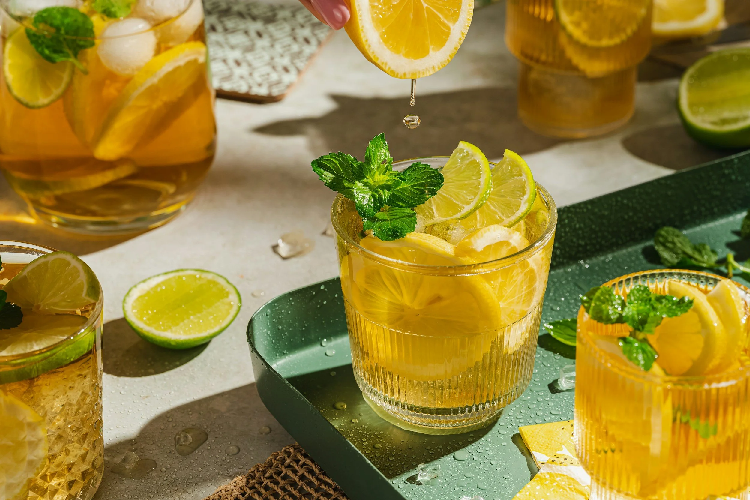 Glasses of iced lemon and mint drinks with lemon slices and fresh mint leaves, surrounded by halved lemons and limes on a bright table.