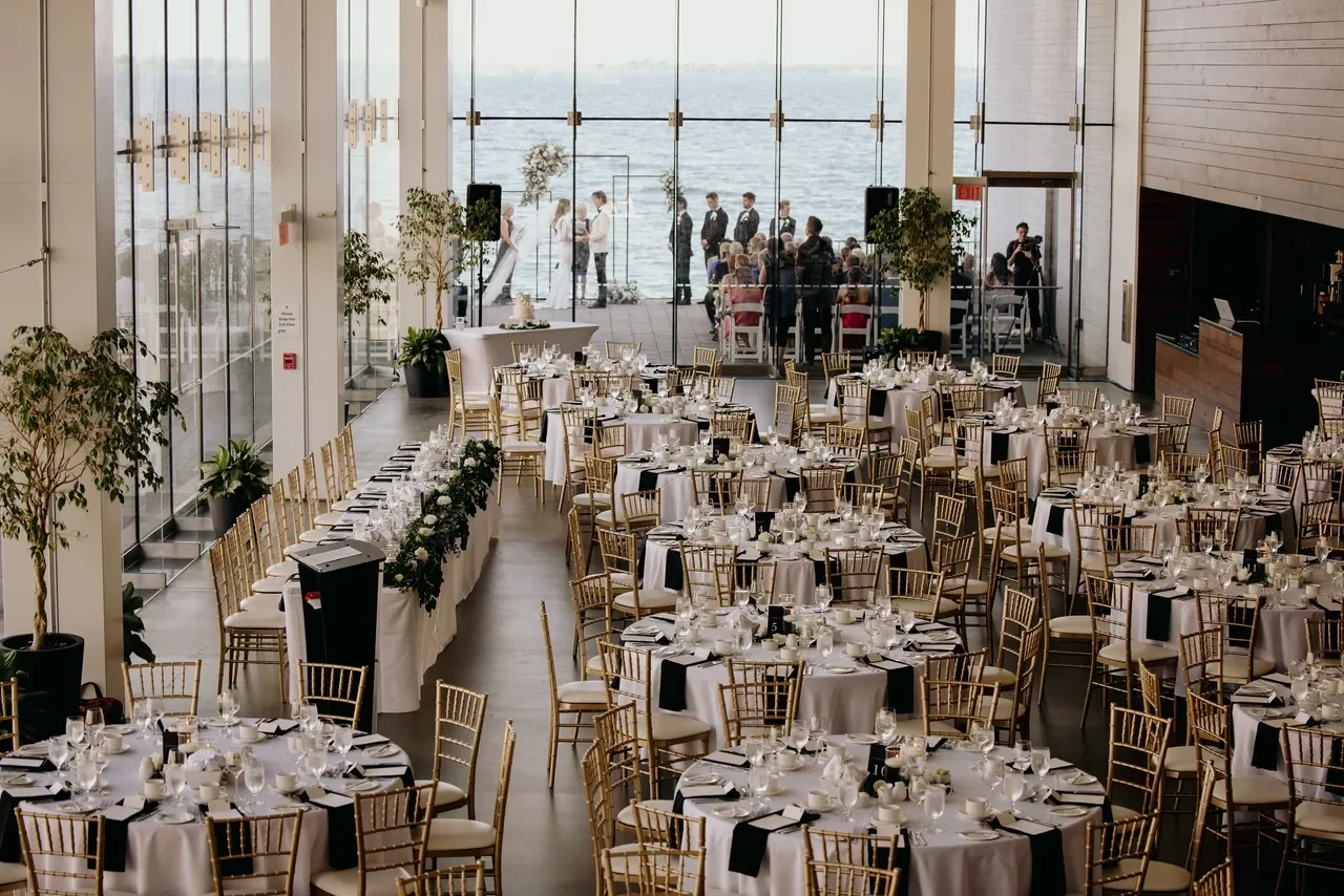 The Isable Bader Centre set with round tables set for a formal event, decorated with white tablecloths, black napkins, and floral centerpieces, featuring large windows overlooking a water body where people are gathered outside.