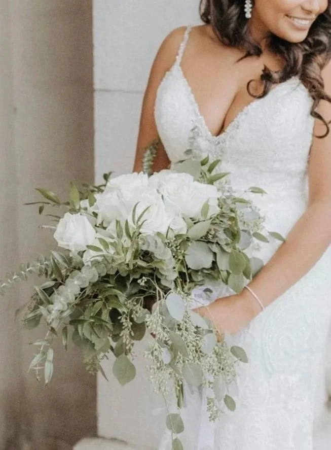 Bride in a spaghetti strap dress with curly black hair carring a large rose and eucalyptus boquet.