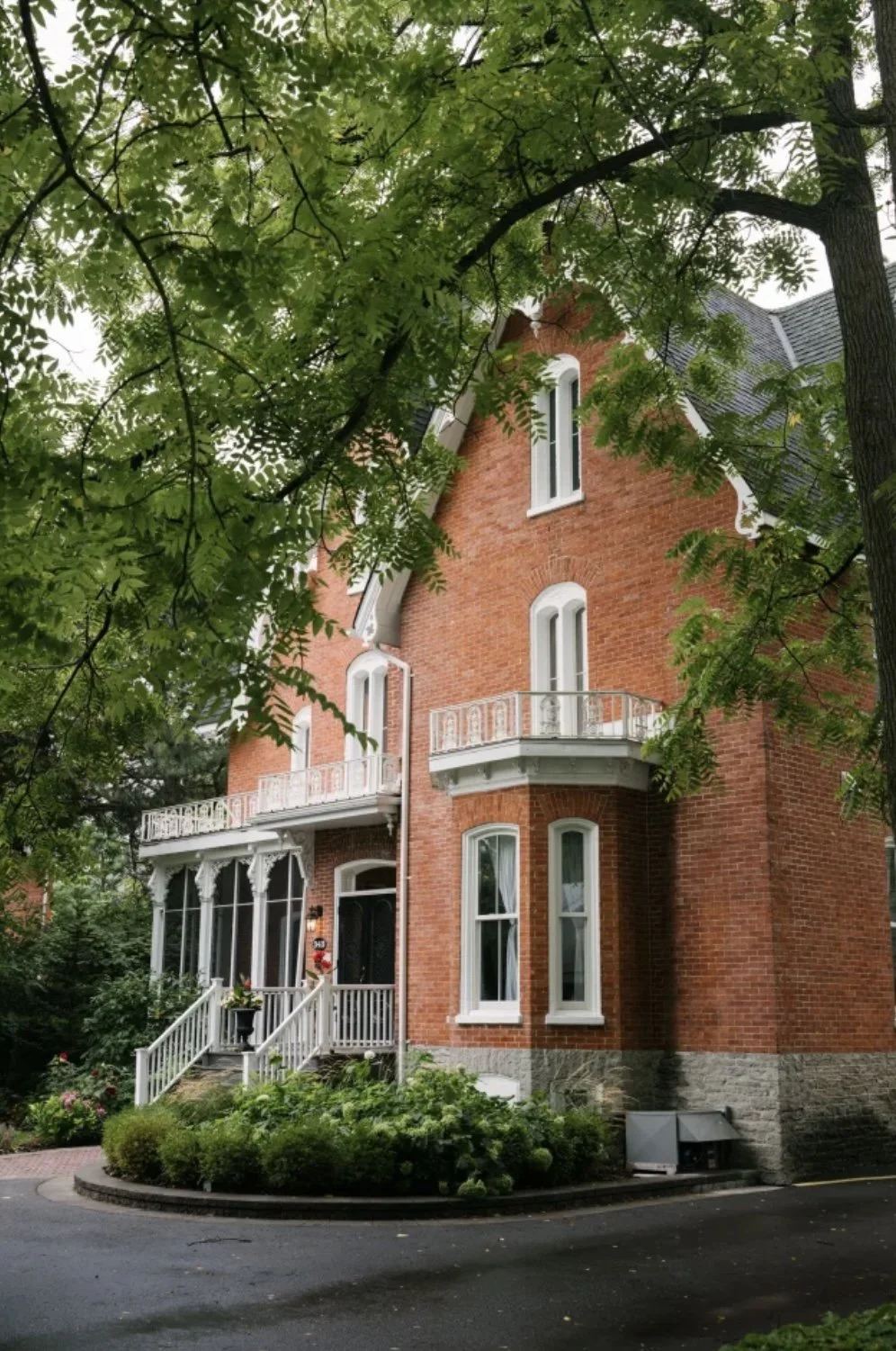 Red brick, victorian house with white arched windows and a front porch surrounded by lush greens.