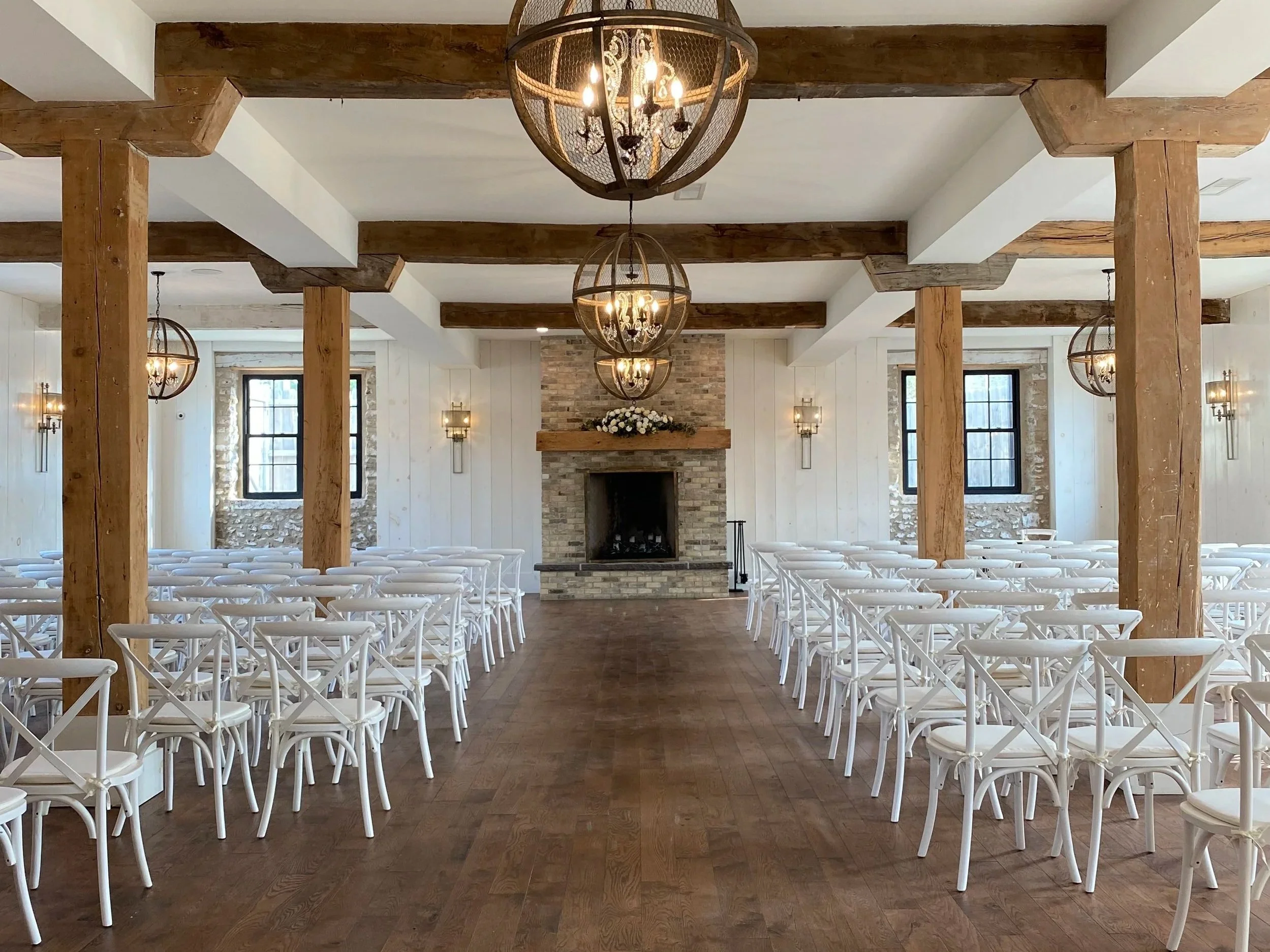 Wedding ceremony infront of a fireplace decorated with white flowers. Wooden beams and chandeliers hang from the ceiling, with large windows on side walls.