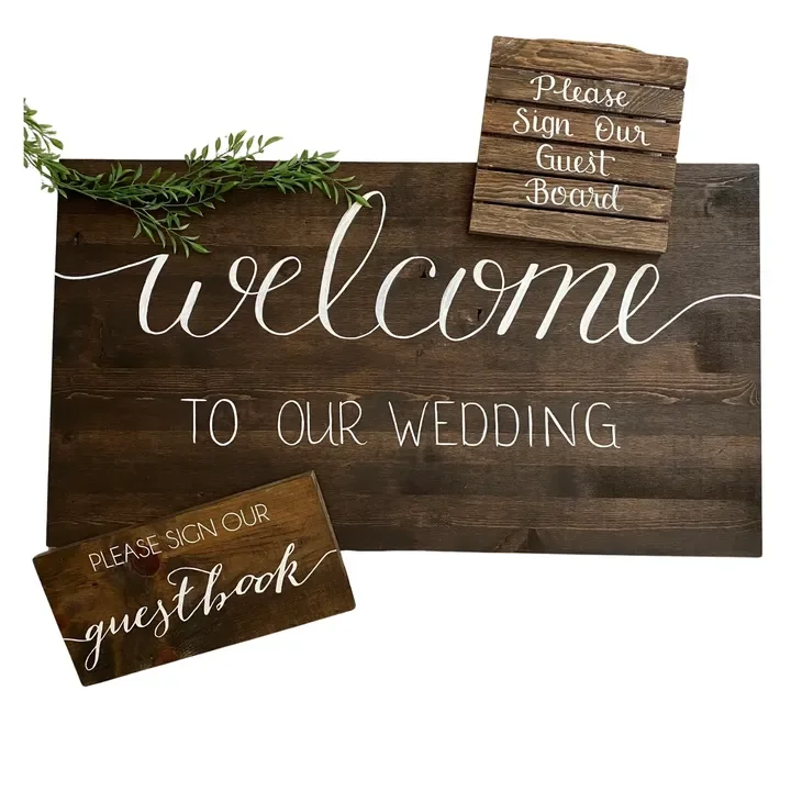 Wooden sign with white cursive and block lettering welcoming guests to a wedding, accompanied by smaller signs requesting guests to sign the guestbook and guestboard, decorated with greenery.
