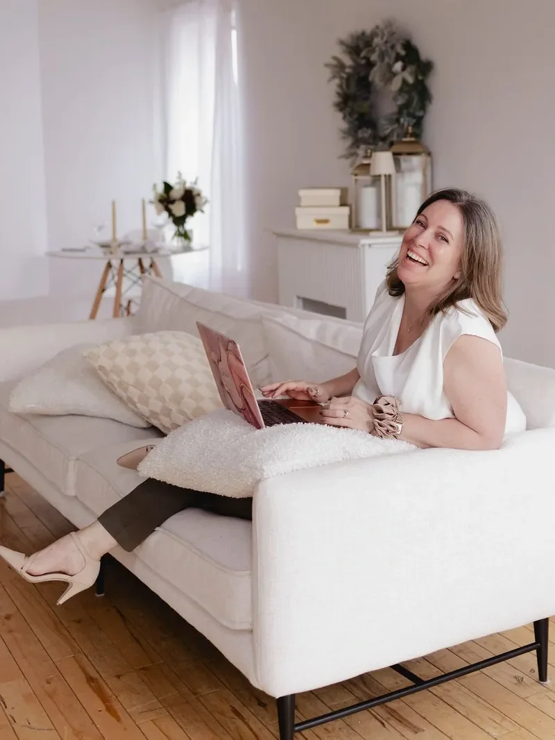 Wedding planner sitting on a white sofa with a laptop, smiling, in a cozy decorated living room with a wreath, candles, and a bouquet of flowers in the background.
