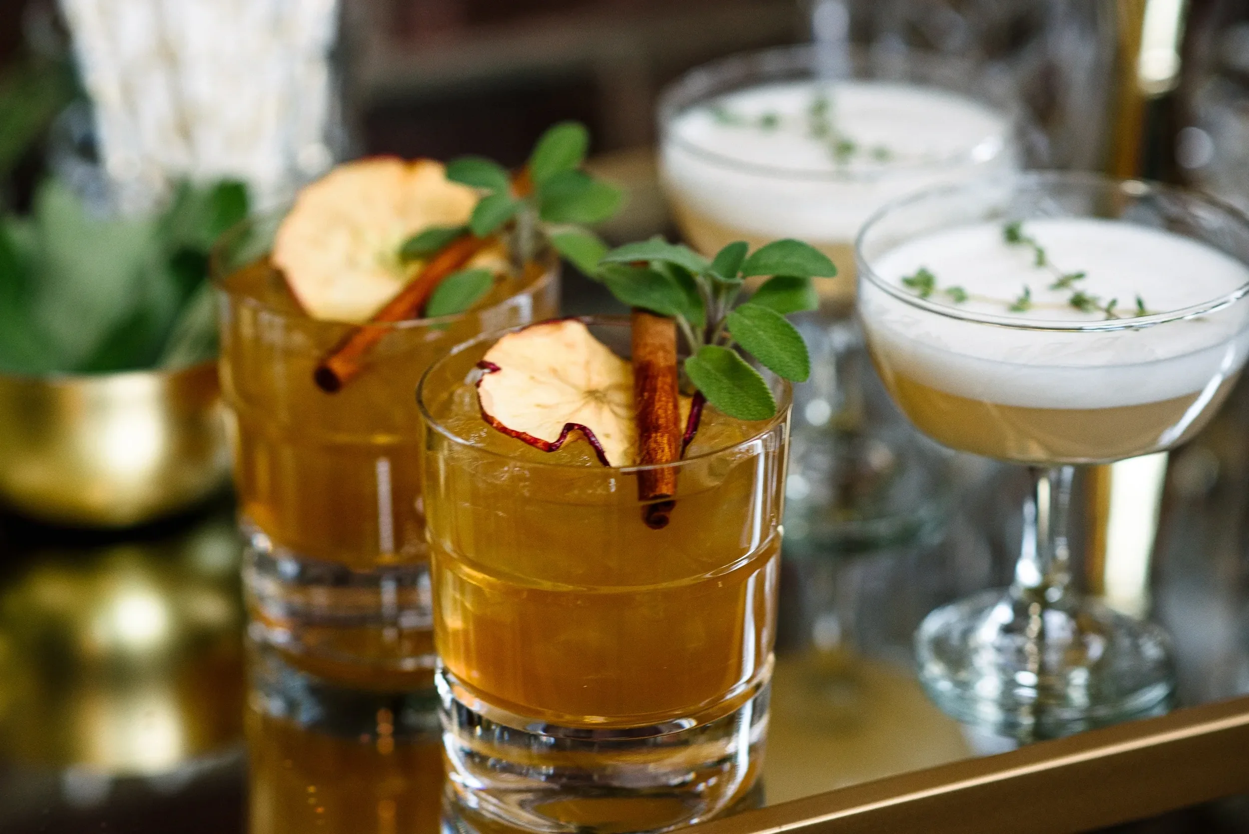 Two glasses with a brown cocktail garnished with apple slices, cinnamon sticks, and mint leaves. In the background, there are two more cocktails with frothy tops and tiny herbs.