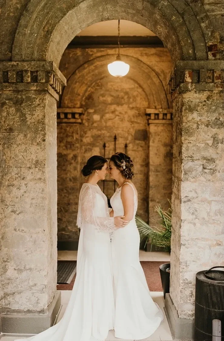 Brides in wedding dresses standing close together inside a stone arch at the Rosemount Inn, touching foreheads and smiling, with warm lighting overhead.