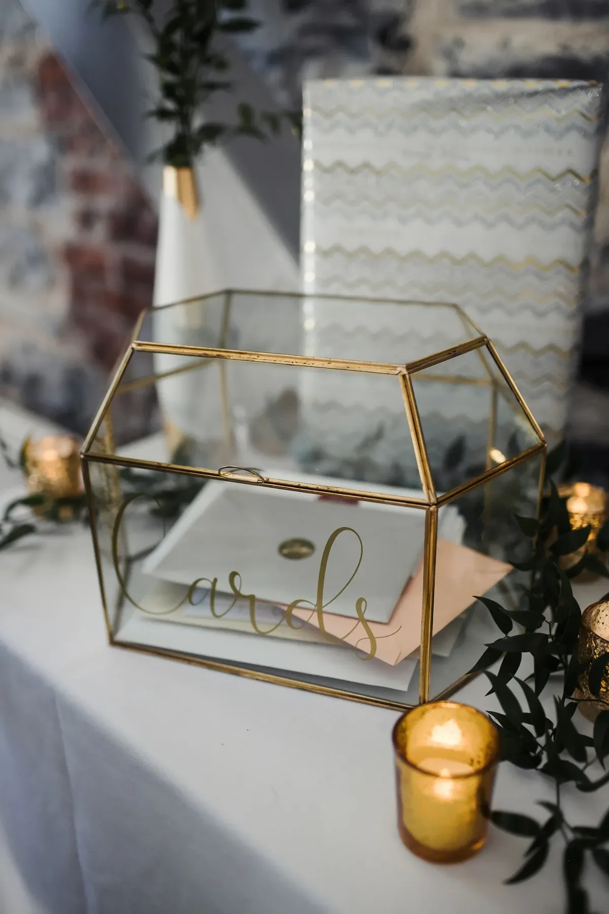 Glass and gold card box with the word 'cards' written on the side, containing folded paper cards inside, surrounded by small candles and greenery, with a decorated backdrop.