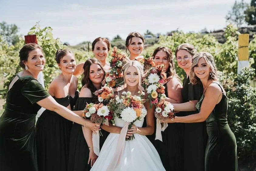A bride with bridesmaids holding bouquets of flowers in an outdoor vineyard setting.