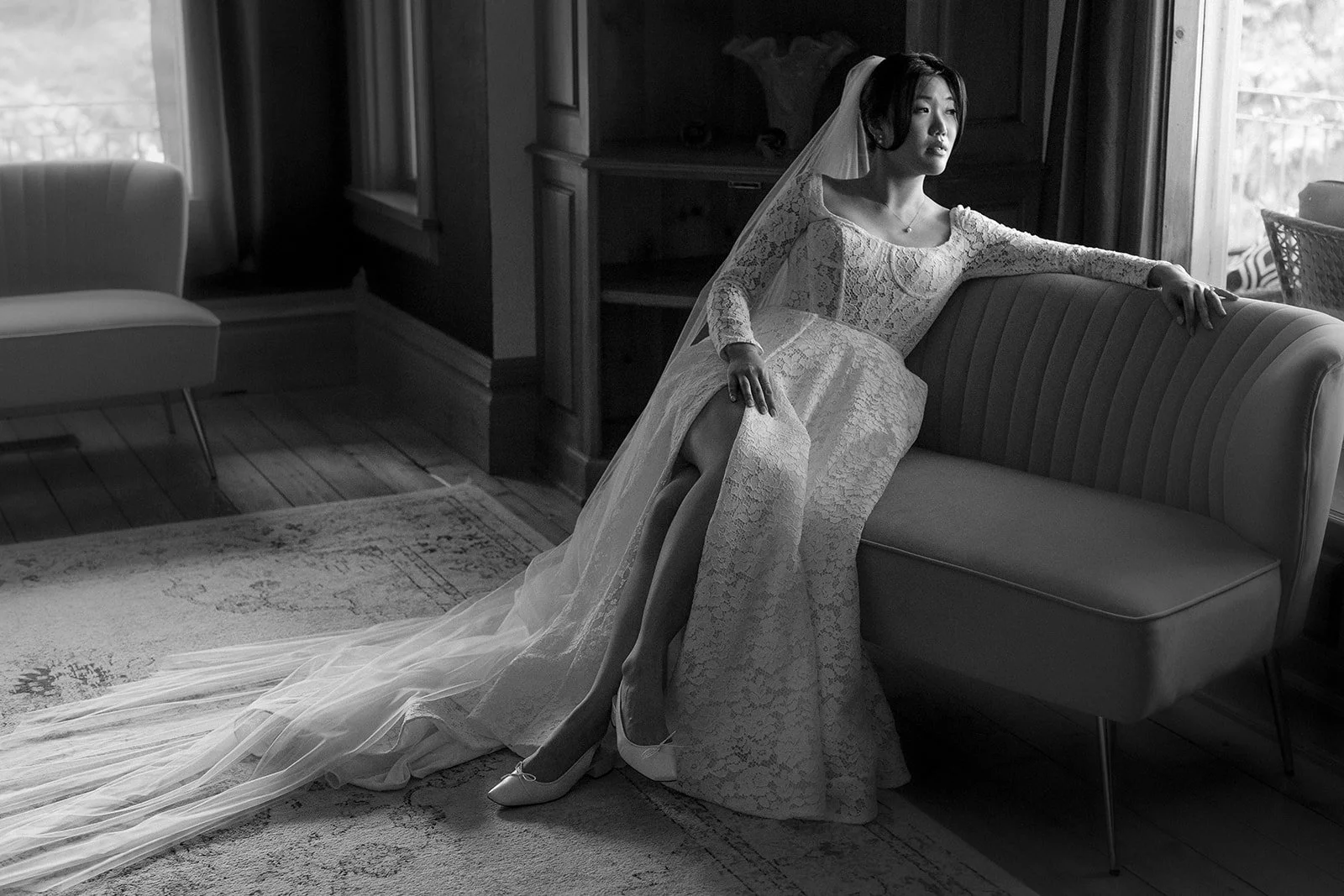 Black and white photo of a woman in a wedding dress and veil sitting on a sofa, with one leg crossed over the other, looking out a window.