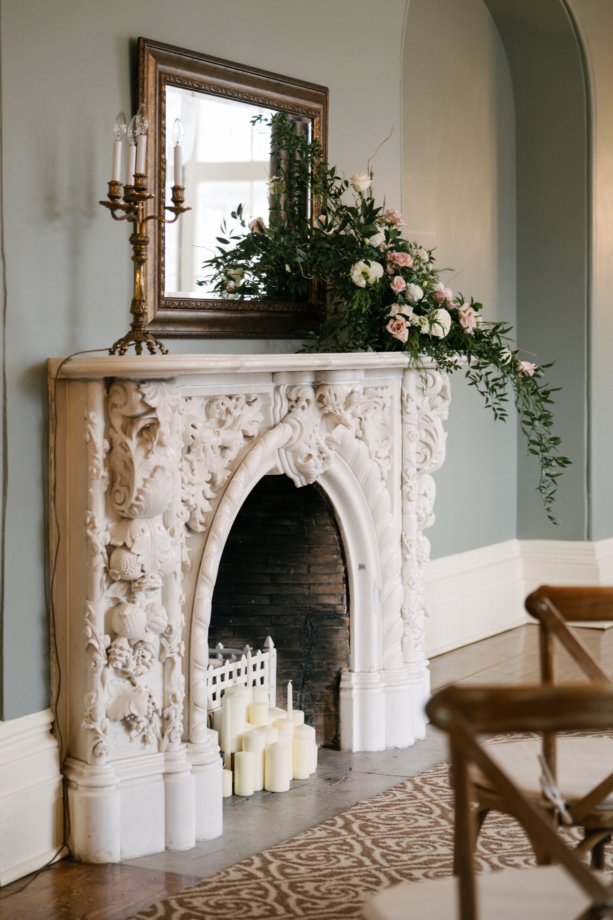 Historic white stone fire place filled with candles and topped with a large floral arrangement and a gold framed mirror.