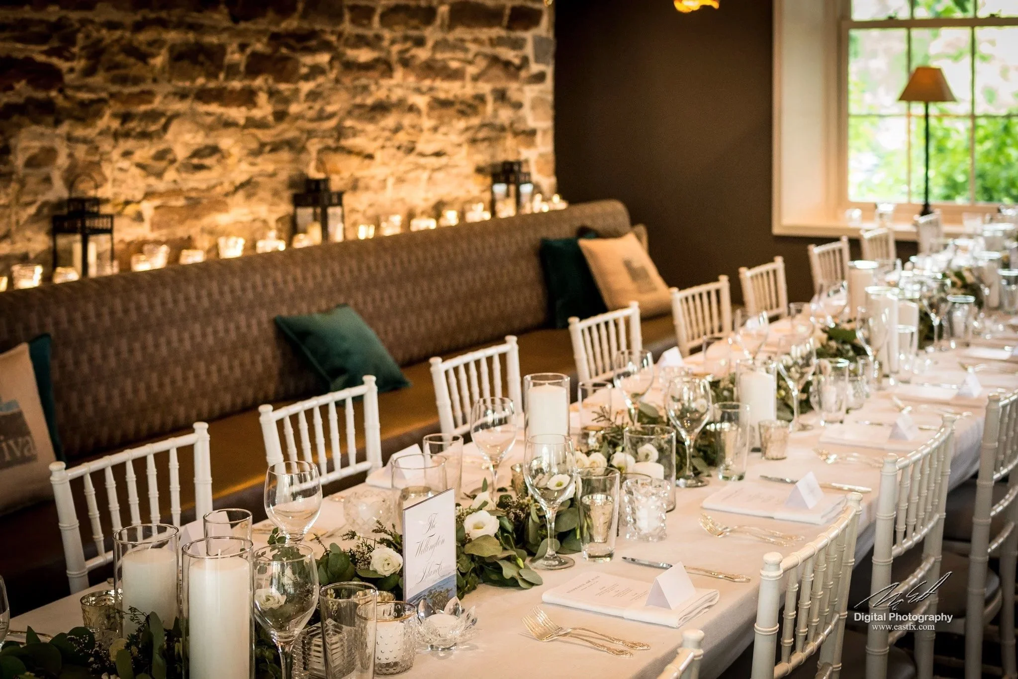 A long banquet table decorated for a wedding reception with white chairs, floral centerpieces with white roses and greenery, candles, and place cards, set against a rustic stone wall and a large window with greenery outside.