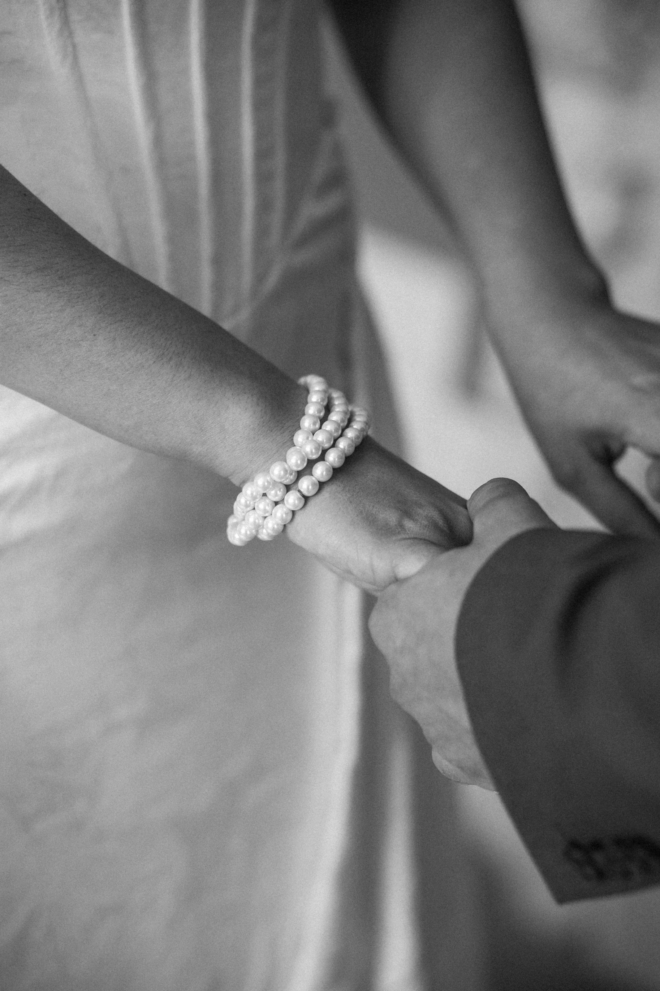 Close-up of two people holding hands, with the person's arm wearing pearl bracelets and a suit, in black and white.