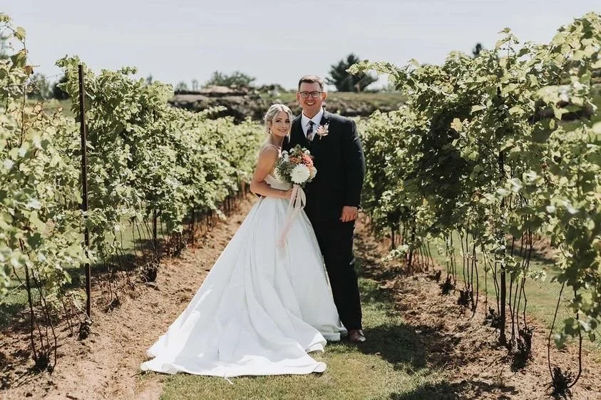 A bride and groom standing in a vineyard on their wedding day, with the bride holding a bouquet of flowers.