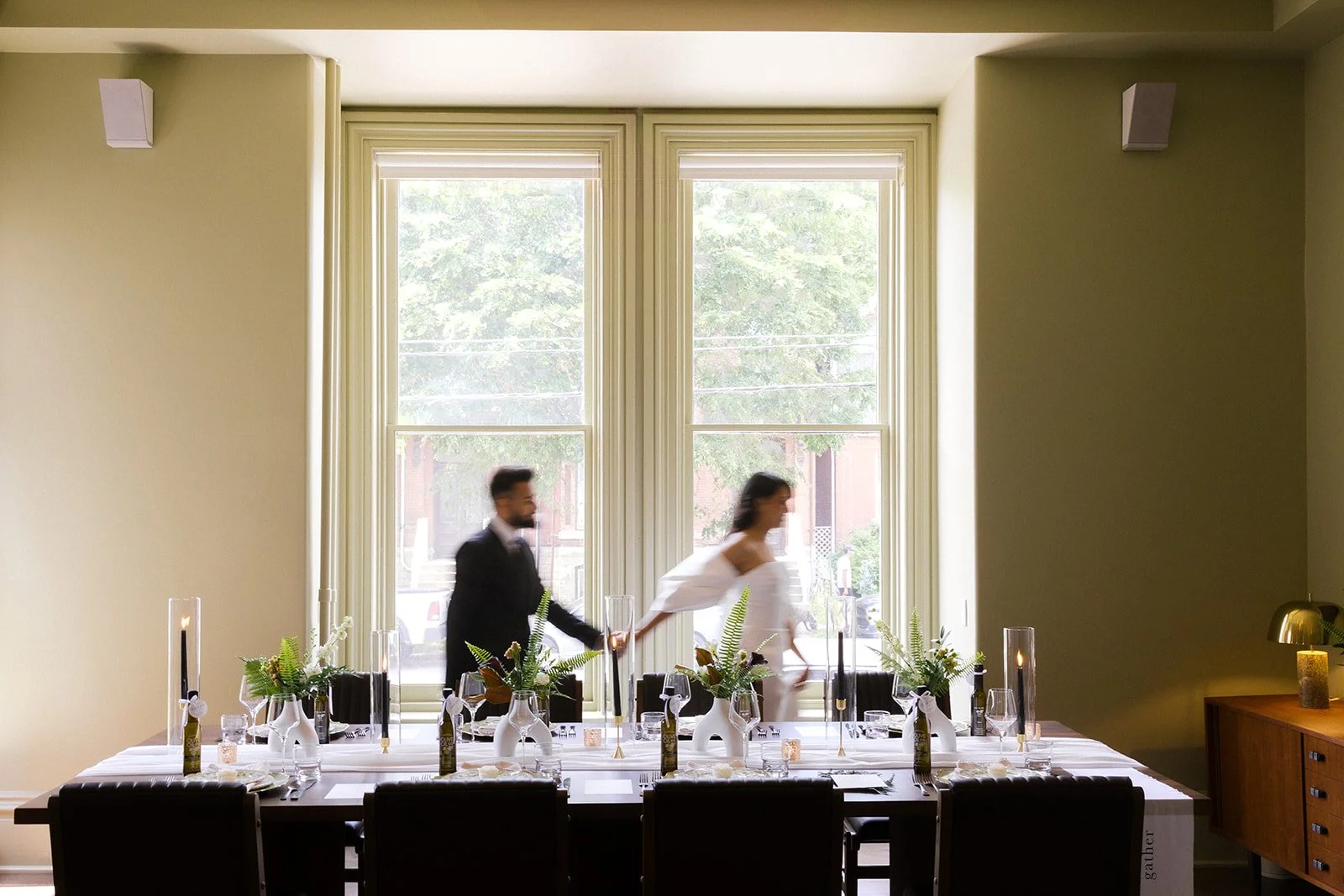 Groom in a tuxedo and a Bride in a white dress, holding hands and walking across a decorated dining table in a room with large window and green walls at the Belevedere Hotel Kingston