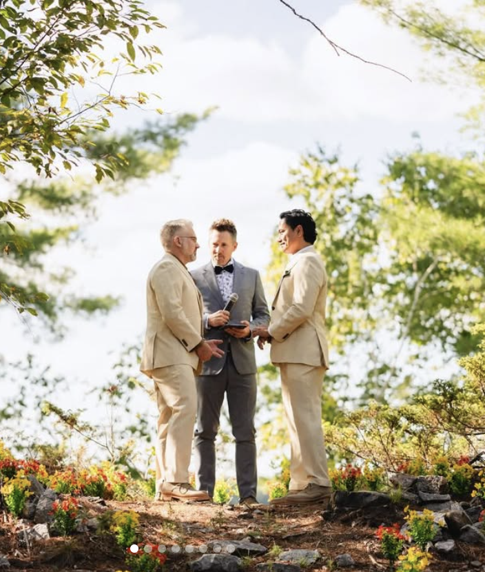Grooms beige suits standing on a rocky ground and holding hands, exchanging wedding vows; officiant in gray suit with bow tie.