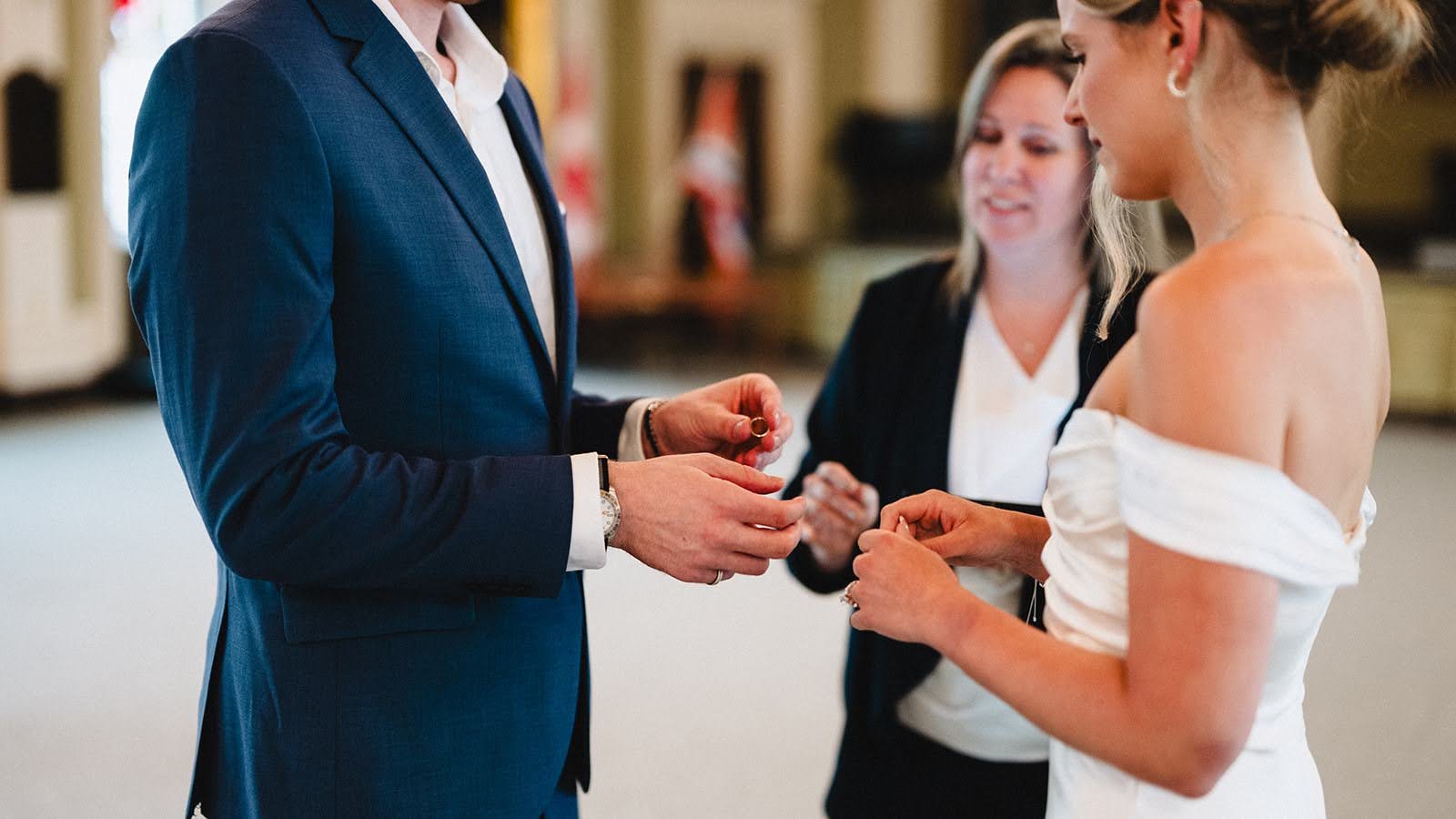 A wedding ceremony with a bride in an off-shoulder white dress and two women, one officiant in a black jacket and one man in a blue suit, exchanging rings.